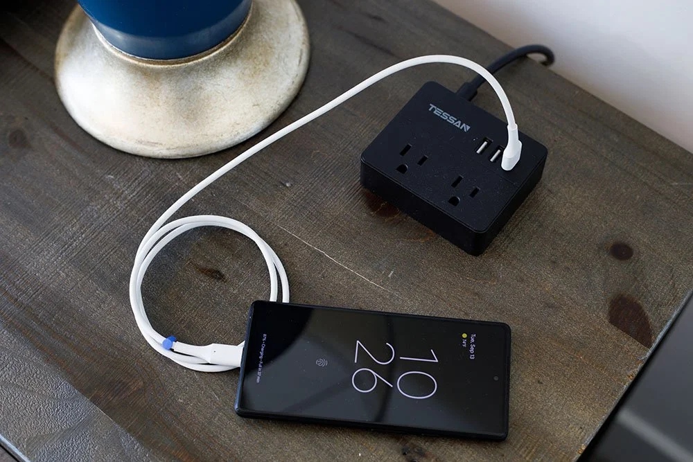 A smartphone connected to a black power strip with a white charging cable, placed on a wooden surface.