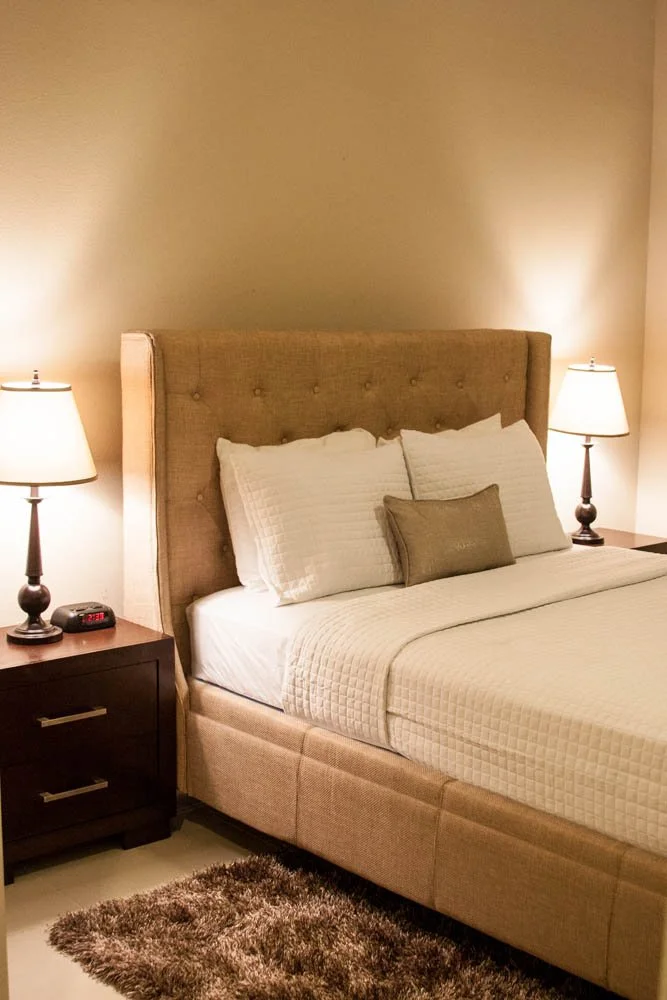 A bedroom with a beige upholstered bed, white bedding, two bedside tables with lamps, available to rent at Furniture Options.