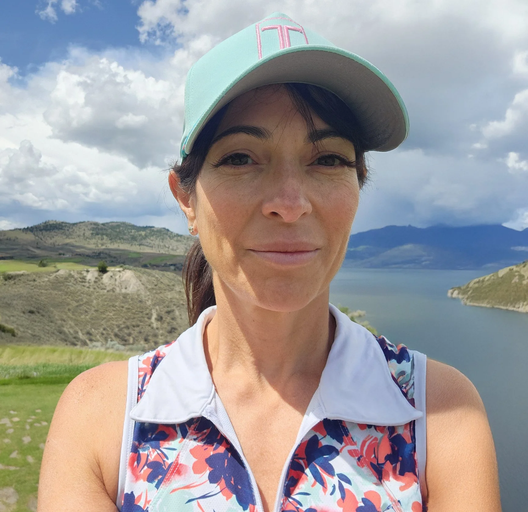 A woman wearing a pastel green cap with pink embroidery, standing outdoors near a body of water with hills and mountains in the background, under a partly cloudy sky.