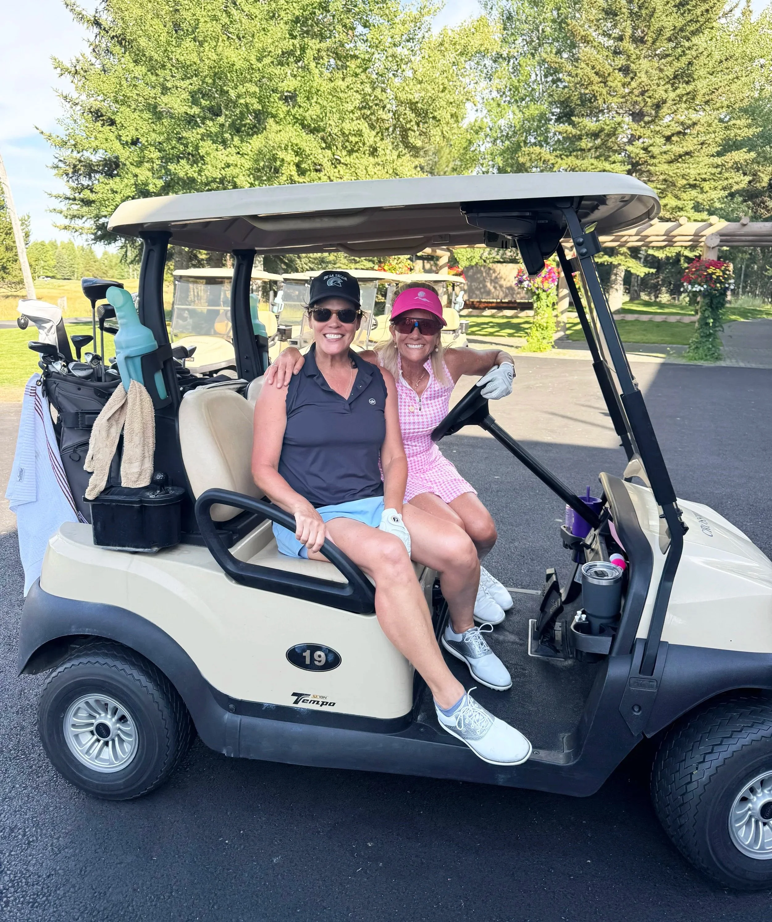 Two women sitting in a golf cart, smiling, with one wearing a pink cap and pink checkered dress, and the other wearing a black cap and sunglasses, on a golf course with trees and flowers in the background.