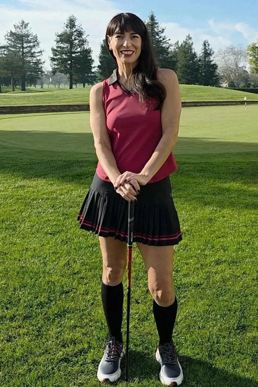 Woman smiling on a golf course, holding a golf club, wearing a sleeveless maroon top, black pleated skirt with red accents, knee-high black socks, and athletic shoes.