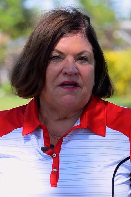 A middle-aged woman with shoulder-length dark brown hair, wearing a red and white collared shirt, standing outdoors with blurred green trees in the background.