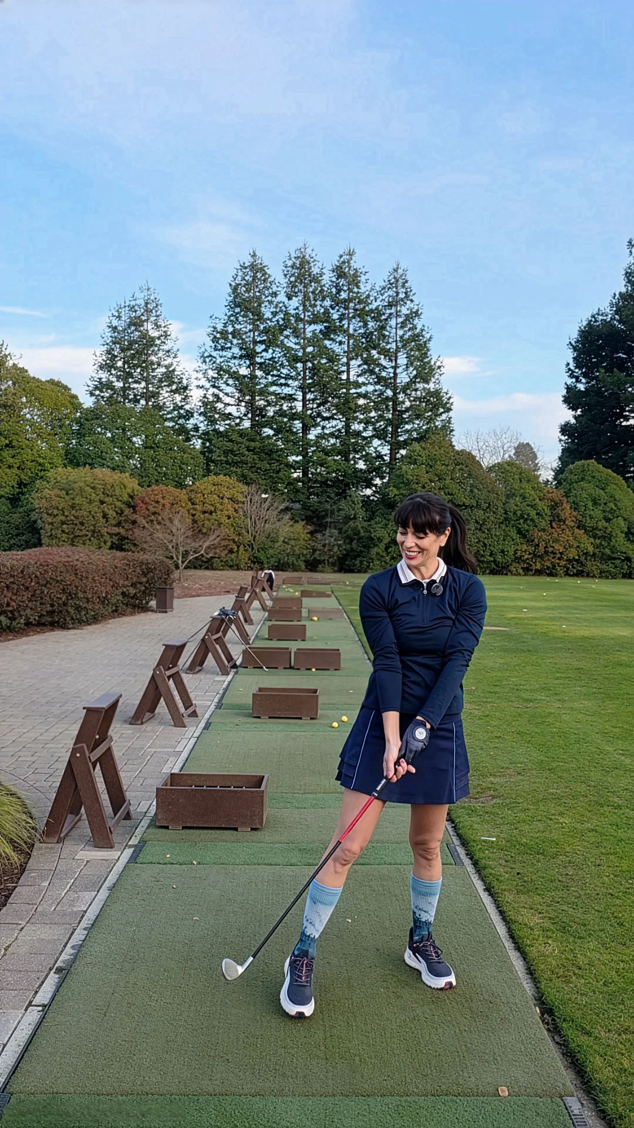 A woman in golf attire practicing her swing at an outdoor driving range surrounded by trees and a clear blue sky.