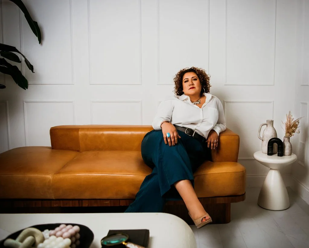 A woman with curly hair sitting on a mustard-colored sofa in a modern room with white paneled walls, wearing a white blouse and dark pants, accessorized with large jewelry and a statement ring, sitting confidently with one leg crossed. A side table with vases and dried plants is beside her.