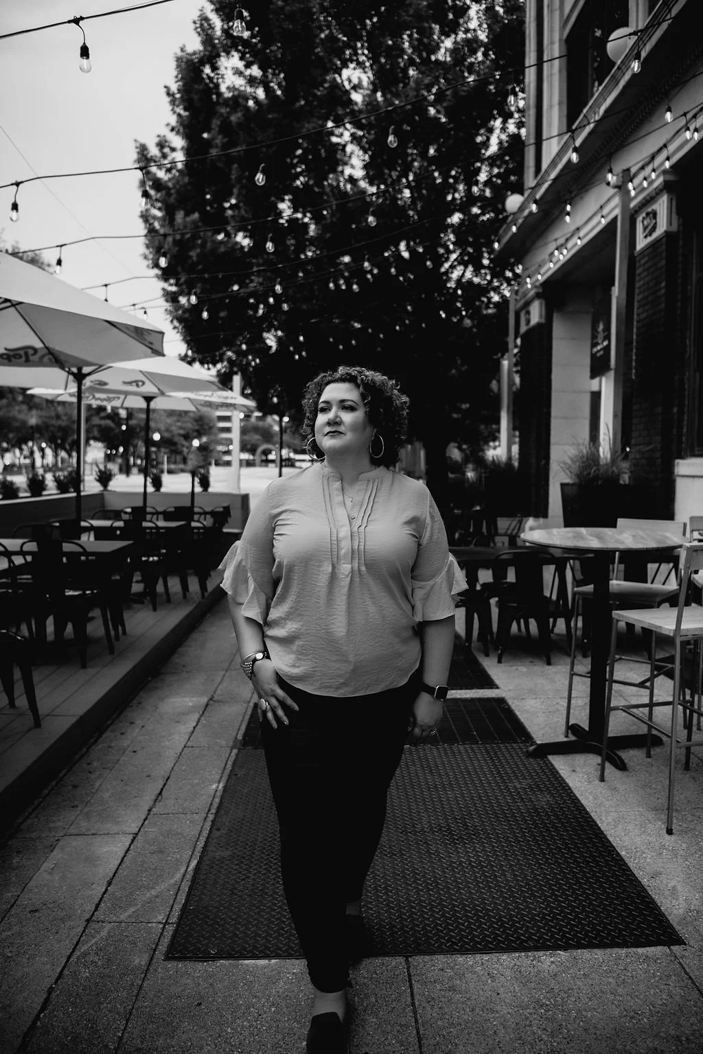 Woman walking outdoors near patio dining area with string lights and umbrellas, black-and-white photo.