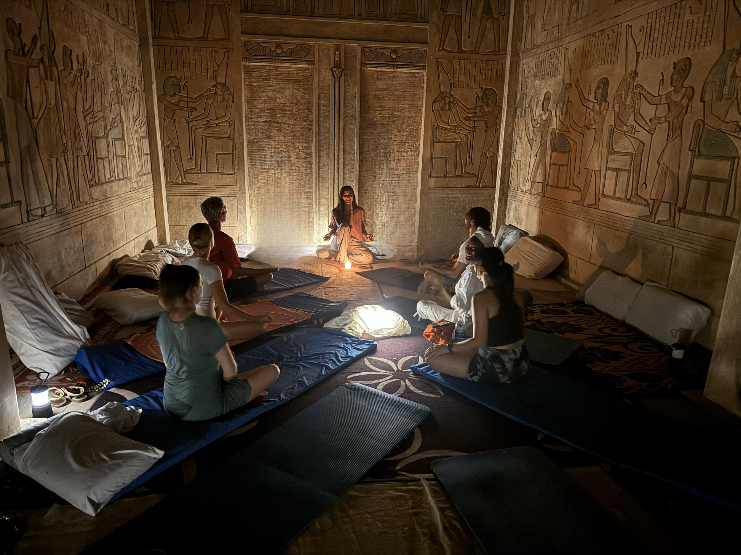 Group of people sitting cross-legged on cushions around a small glowing lamp in a room with Egyptian-style wall art and hieroglyphs, during a meditation or spiritual session.