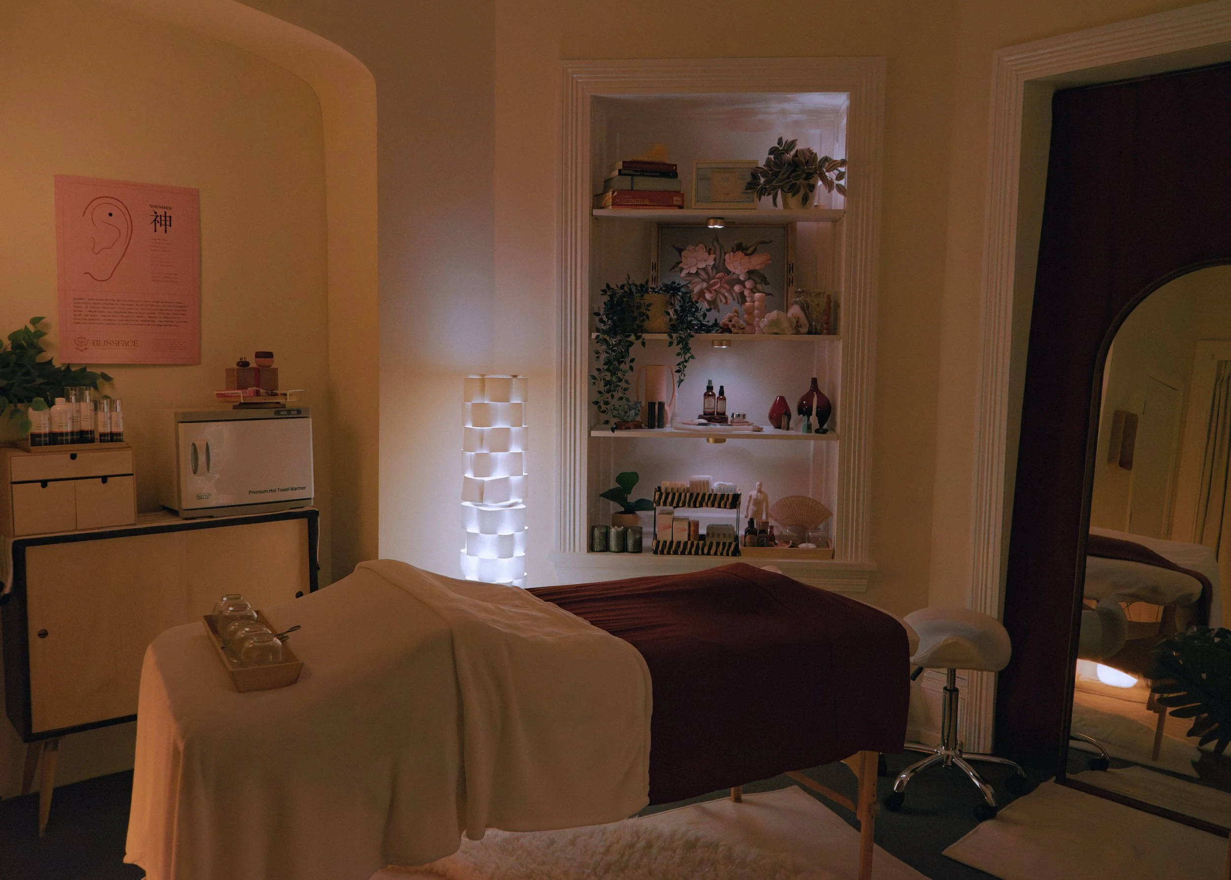 A massage therapy room with a massage table covered in white and burgundy sheets, a tray with massage oils, a tall white lamp, a built-in shelf with decor and skincare products, a mirror, and a folding screen reflecting the room.
