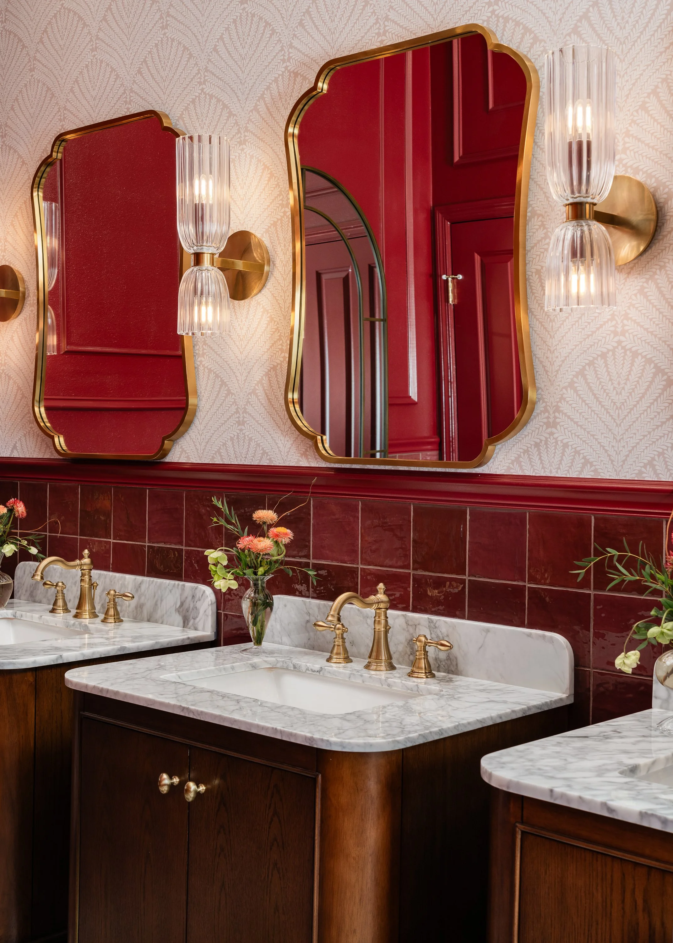 Commercial restroom design. A bathroom vanity with marble countertops, brass faucets, and ornate gold-framed mirrors reflecting a red door. White wallpaper with a subtle pattern adorns the wall above the mirrors.