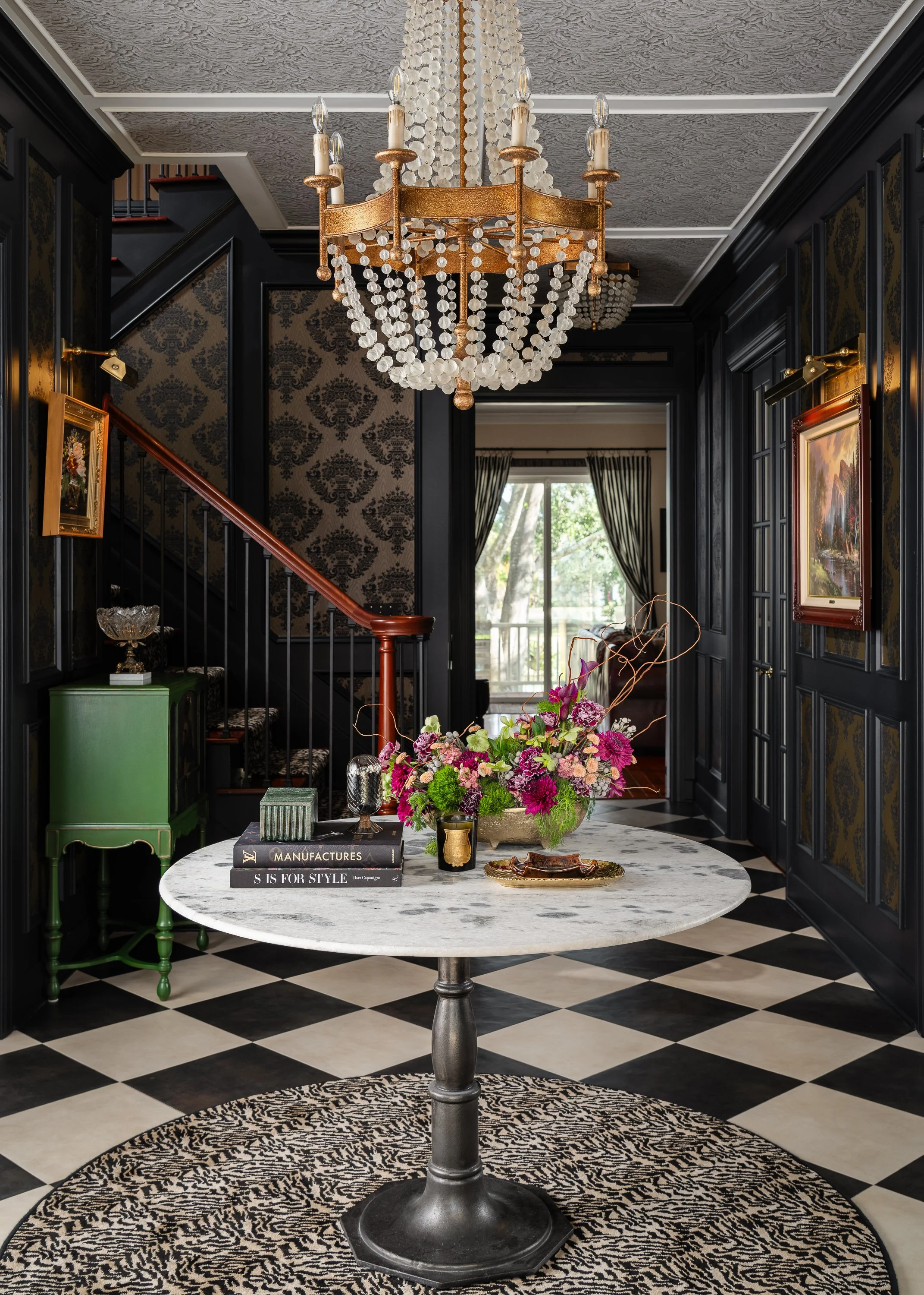 Elegant interior with black paneled walls, a marble table with floral arrangement, books, and Art Deco decorative items, black and white checkered floor, and a chandelier hanging from a textured ceiling.