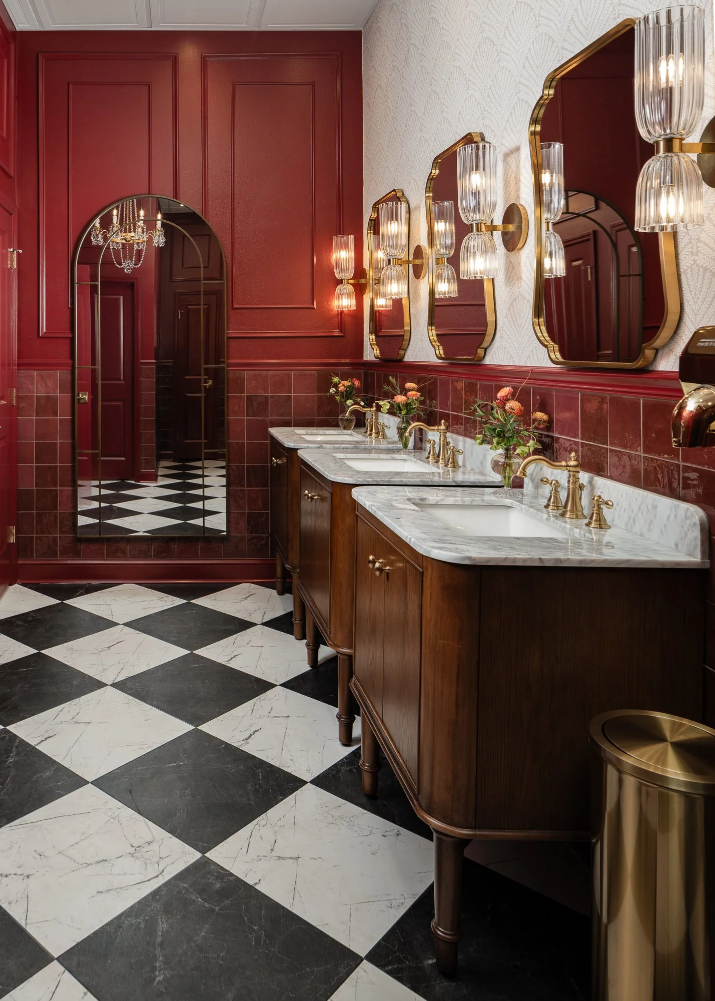 A commercial restroom design with red walls, a checkered floor, wooden vanities with marble countertops, and ornate mirrors with sconces above each sink.