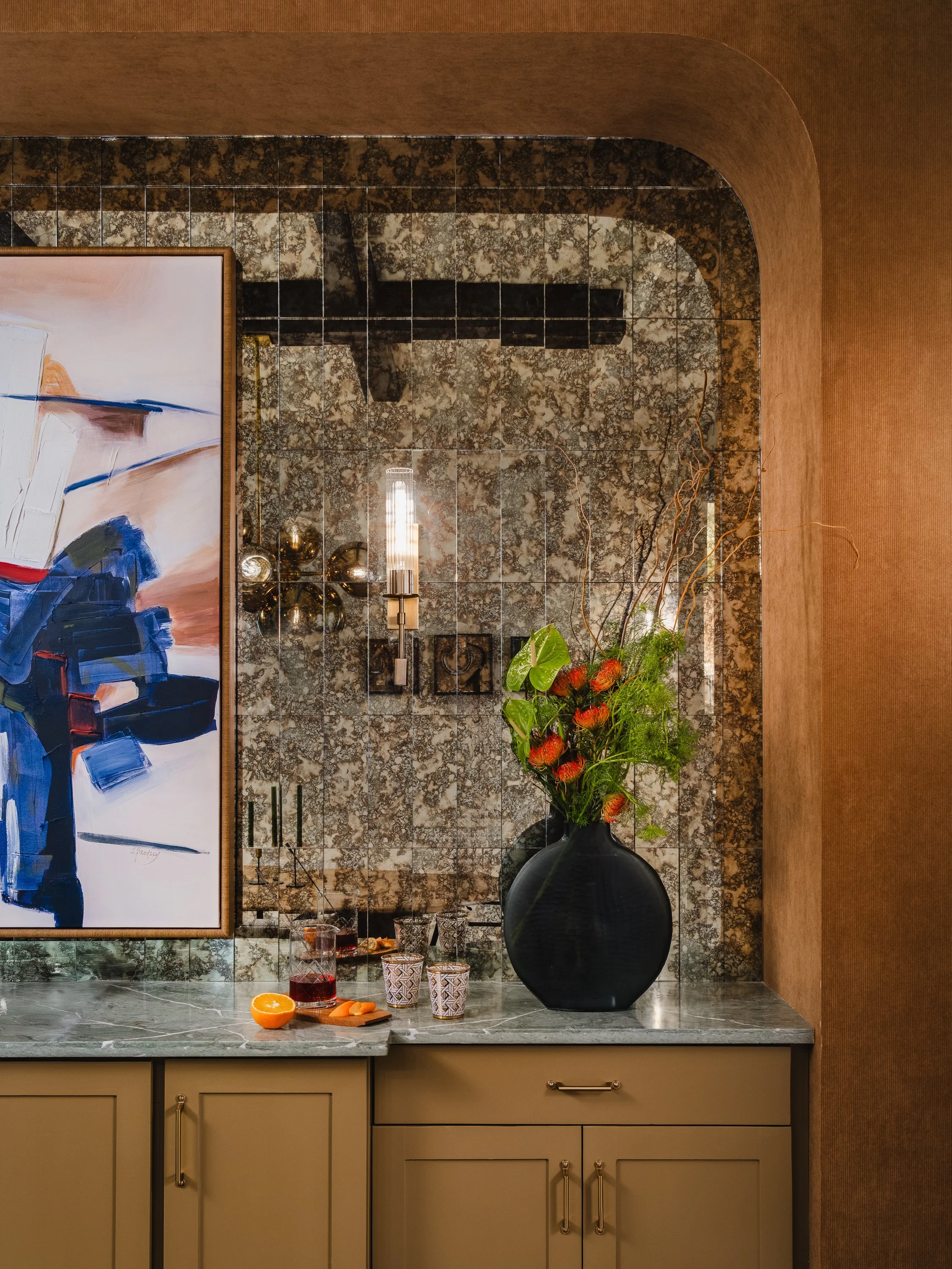 A modern bar area with a large abstract painting, a vase of orange flowers, and drinks on a marble countertop.
