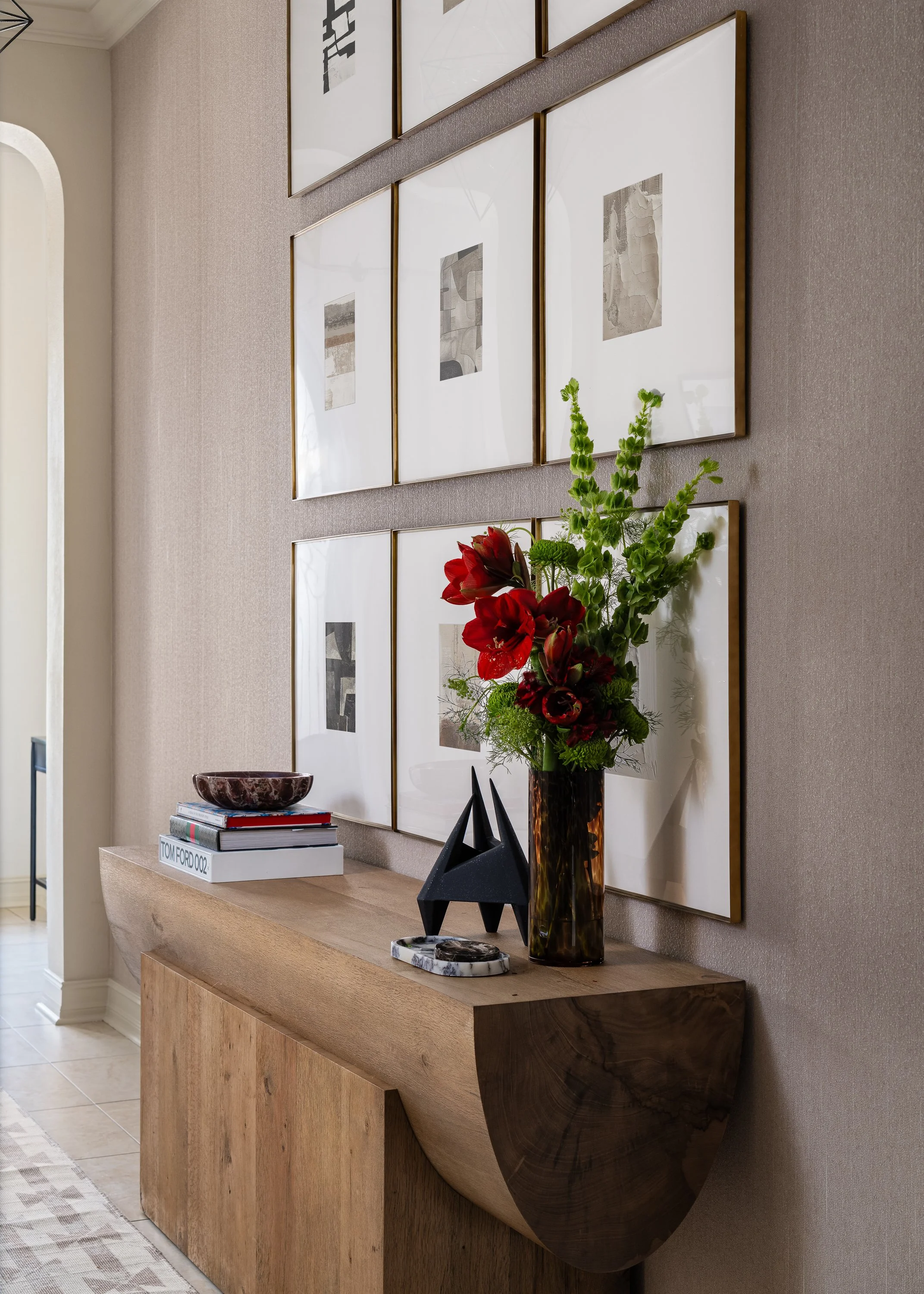 A wooden console table displays a stack of books topped with a decorative bowl, a black sculpture, and a vase of red flowers and greenery against a wall adorned with framed artwork.