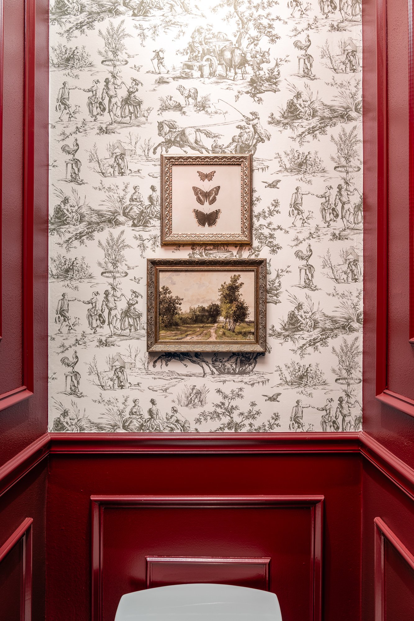 A commercial restroom design wall with toile de Jouy wallpaper and dark red wainscoting features two framed artworks: a shadow box with three dark butterflies and a landscape painting.