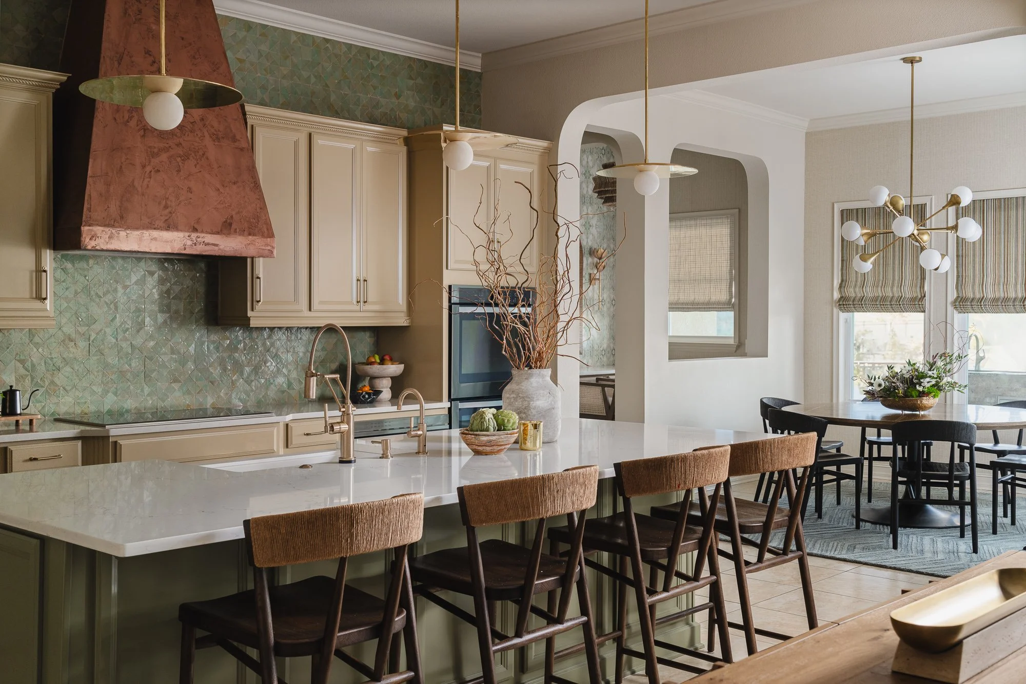Modern kitchen with white cabinetry, a large island with a white marble countertop, brown upholstered bar stools, a decorative vase with branches, and pendant lighting fixtures in gold and white.