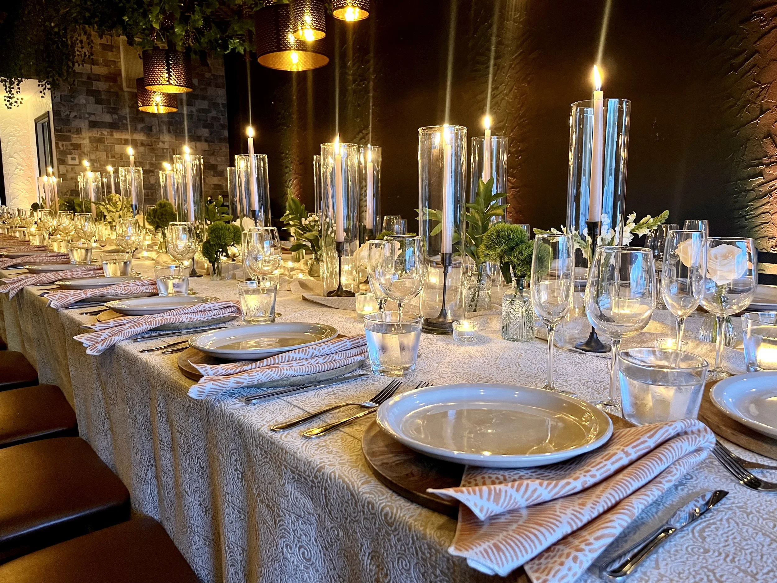 Bespoke wedding tablescape with white textured tablecloth, coral-colored patterned napkins, candles in hurricane candle holders, golden pendant lights, greenery, gleaming white plates, and natural wood charger plates.
