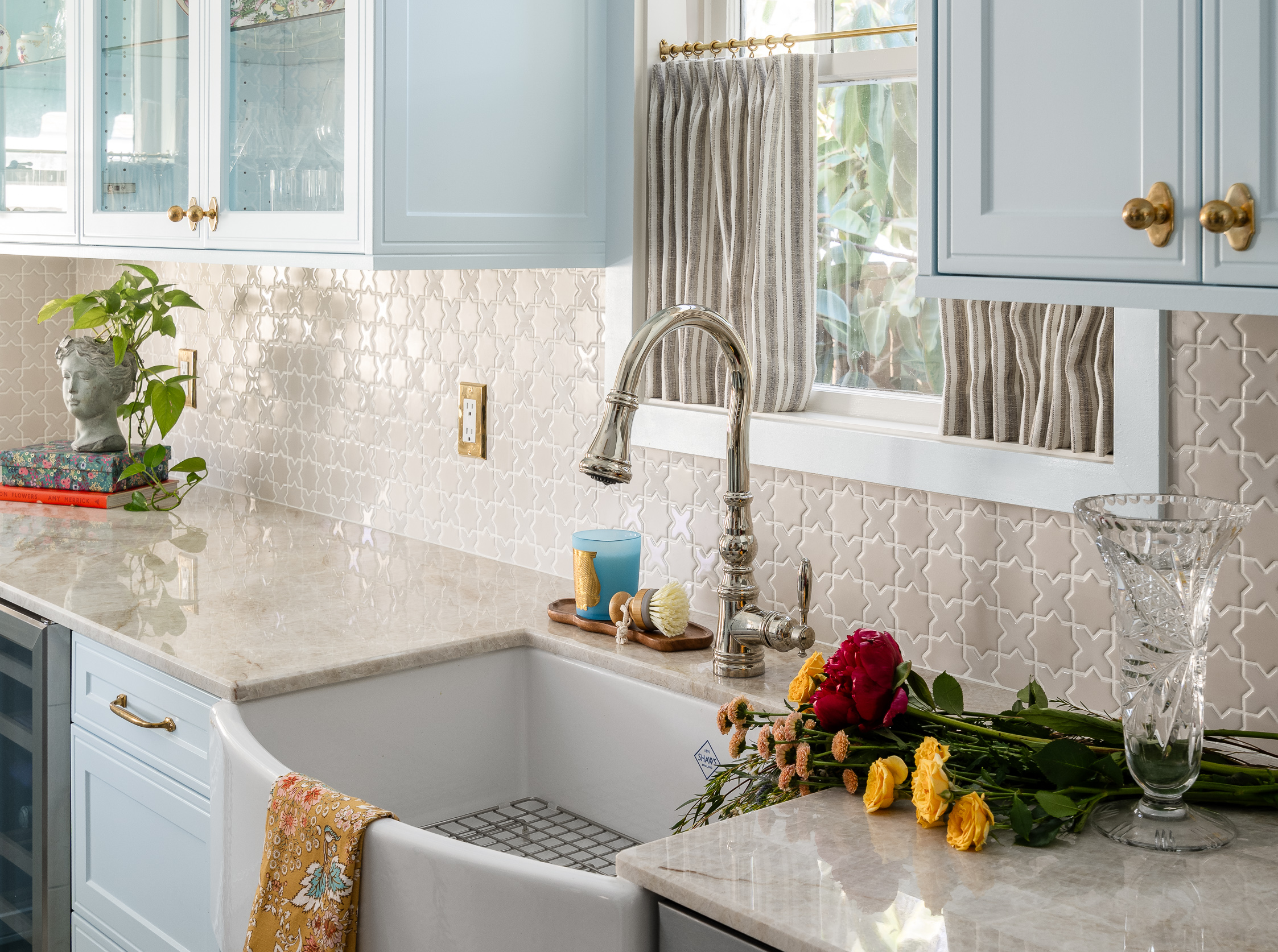 A kitchen sink area with a white farmhouse sink, a chrome faucet, and a backsplash of small, light-colored star-shaped tiles. A bouquet of red and yellow flowers rests on the countertop next to a crystal vase.