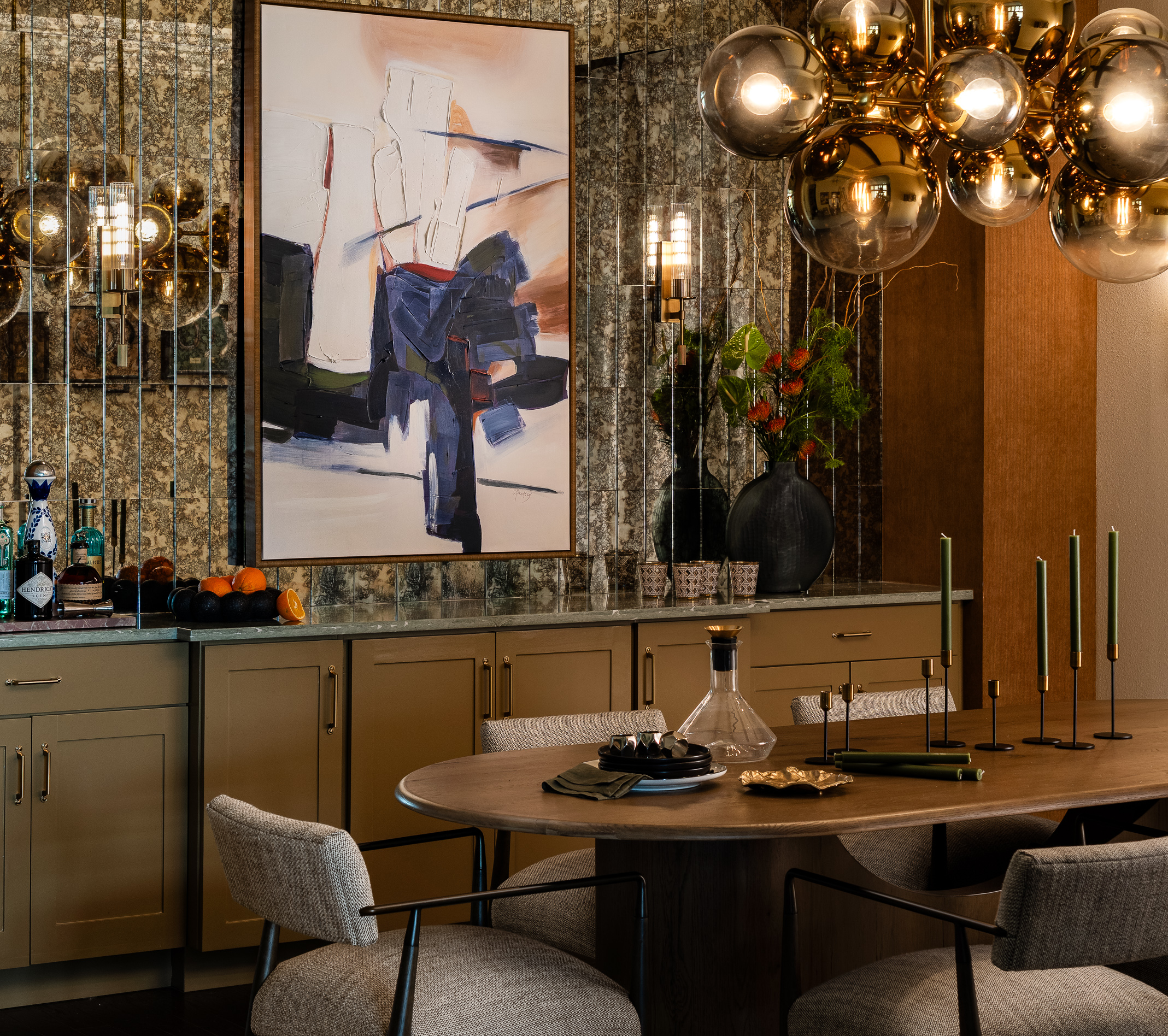 Warm-lit modern dining room with gold globe pendant lights, a large abstract framed painting on a marble-pattern wall, and a countertop set with utensils, bottles, fruit, and glassware.