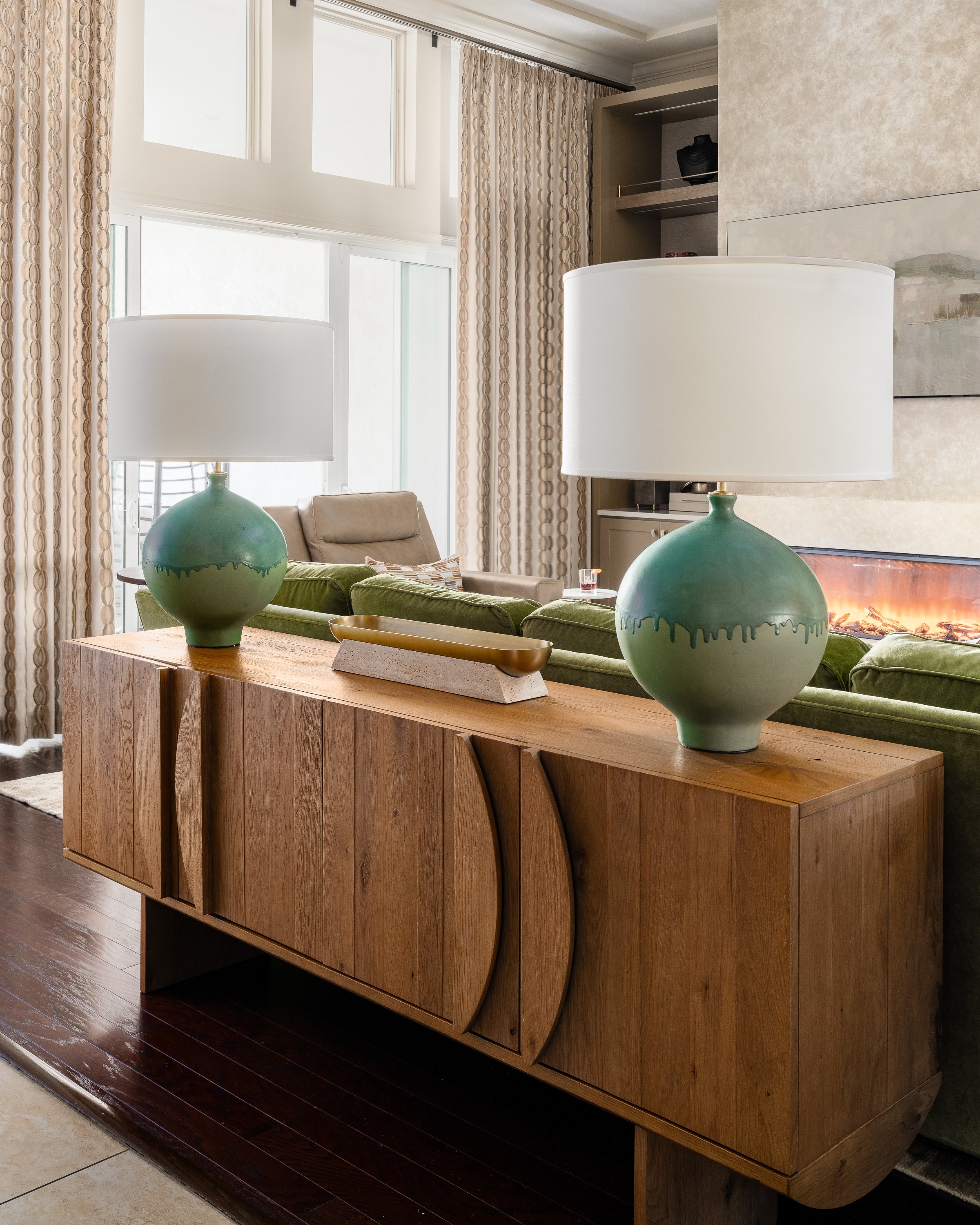 A wooden sideboard with decorative carved doors holds two teal lamps with white shades. A green sofa and a fireplace are visible in the background.