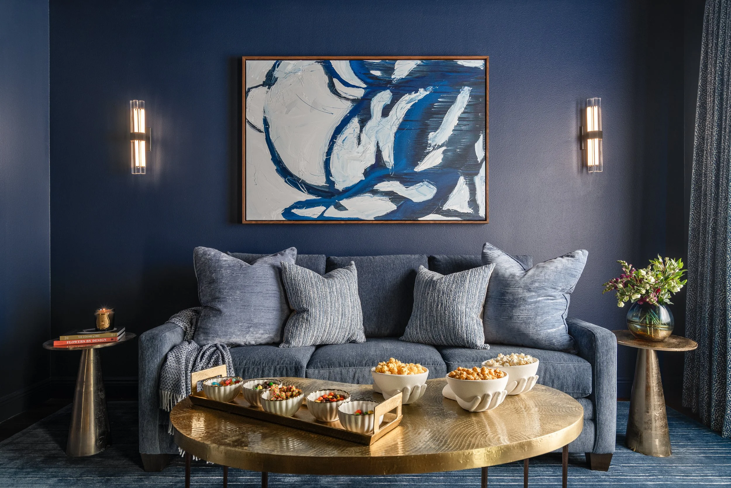 A stylish guest room/media room featuring a blue velvet sofa with light blue throw pillows, a round gold coffee table holding bowls of popcorn and other snacks, and an abstract blue and white painting hanging above the sofa on a dark blue wall.