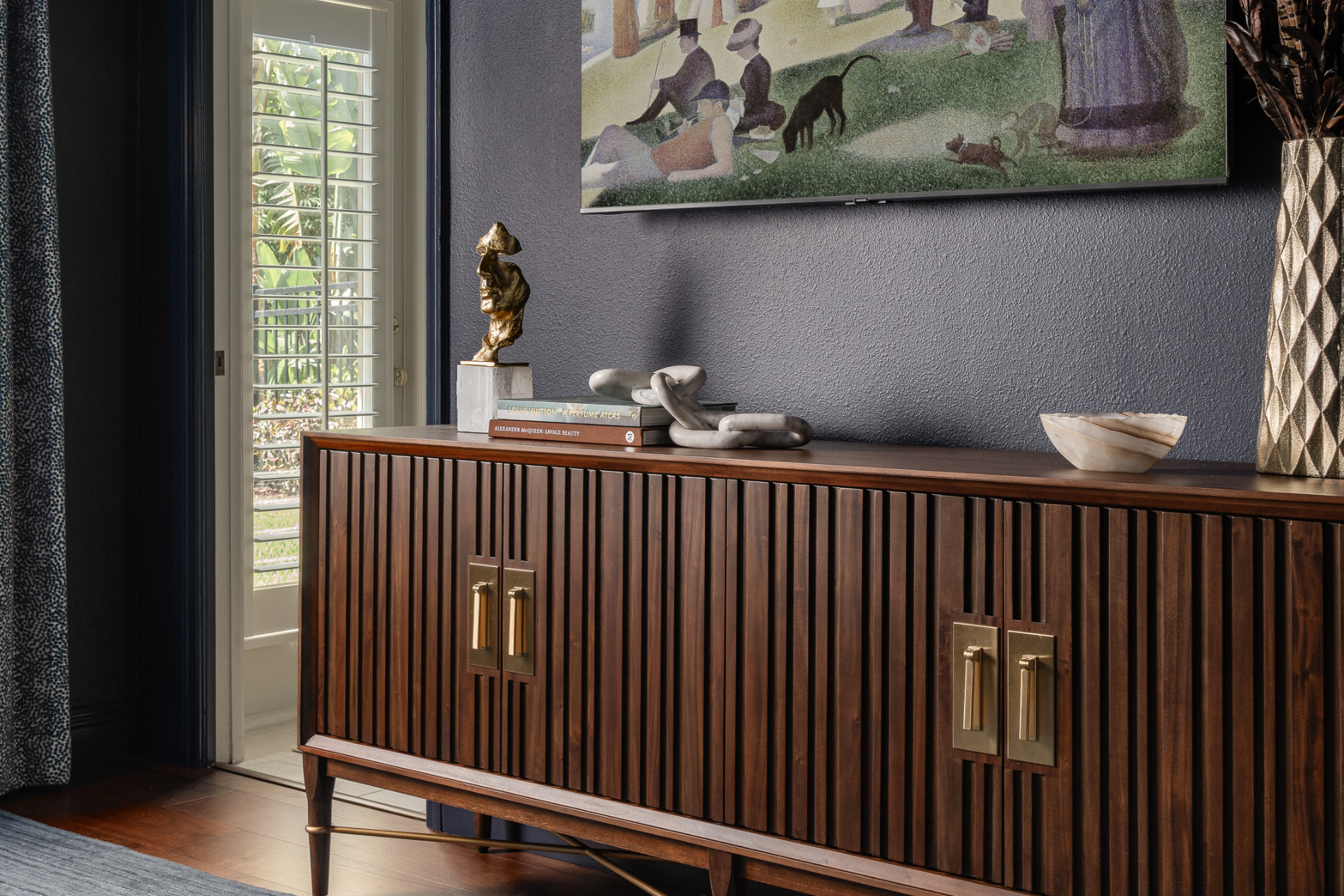 A dark wood credenza with brass handles, in front of a dark painted wall with a colorful impressionist painting. The credenza has carefully curated decor that reflects the owners’ personalities.