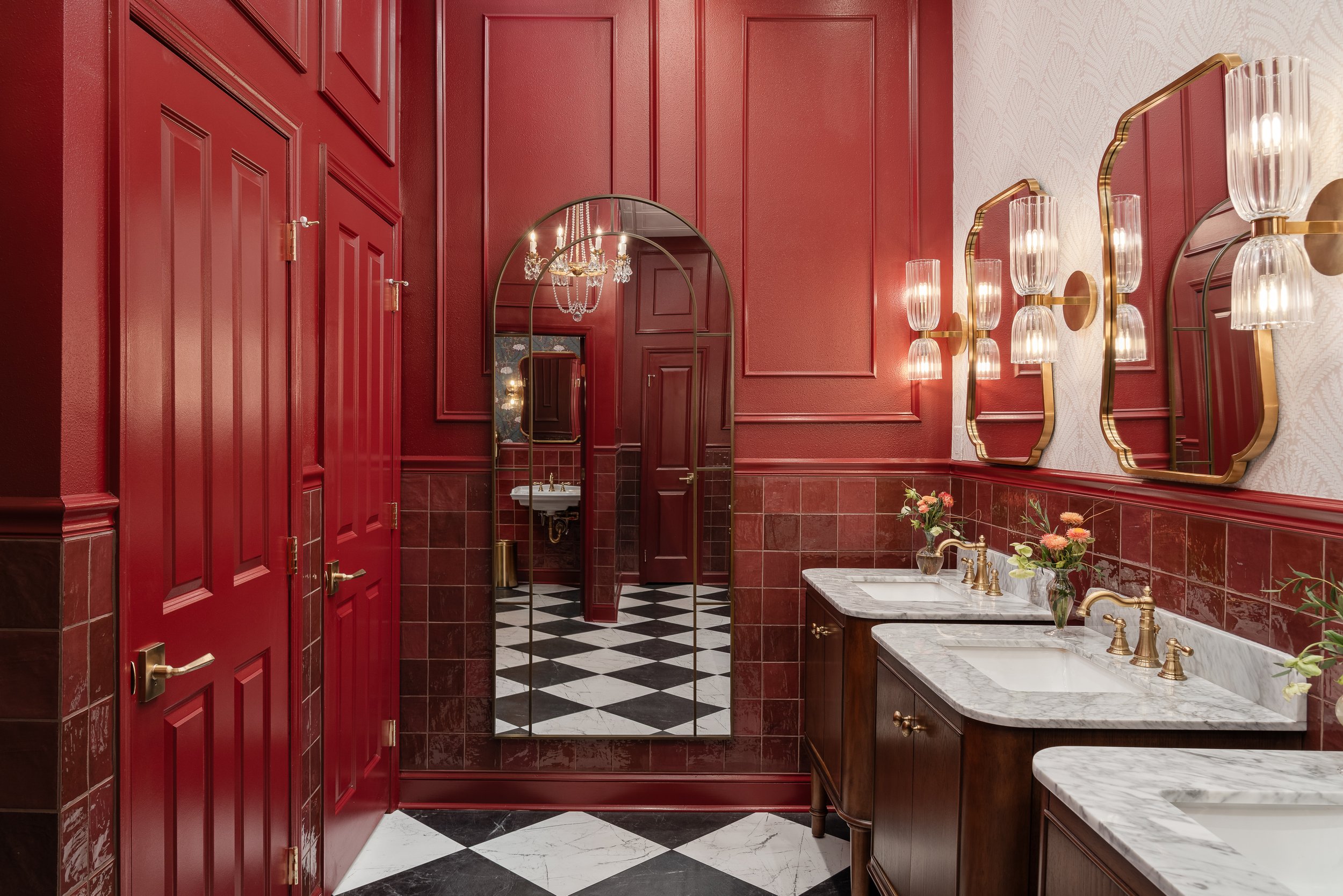 A luxurious red commercial restroom design with a checkered black and white floor, featuring multiple sinks with marble countertops and brass fixtures, ornate mirrors, and decorative wall sconces.