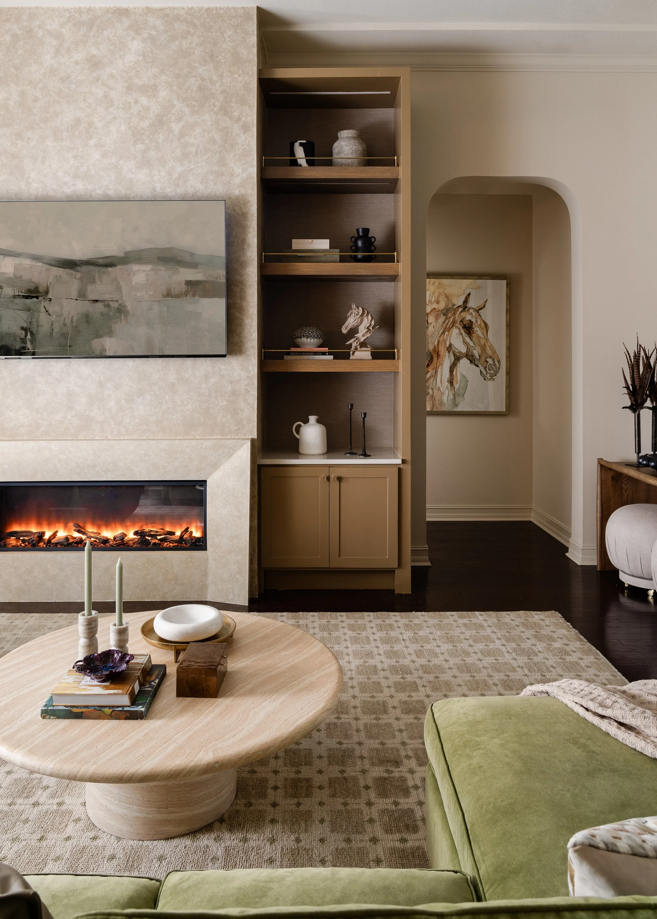 A modern living room featuring a fireplace with a large abstract landscape painting above it, built-in shelving with decorative items, a round coffee table with books and candles, and a green sofa.
