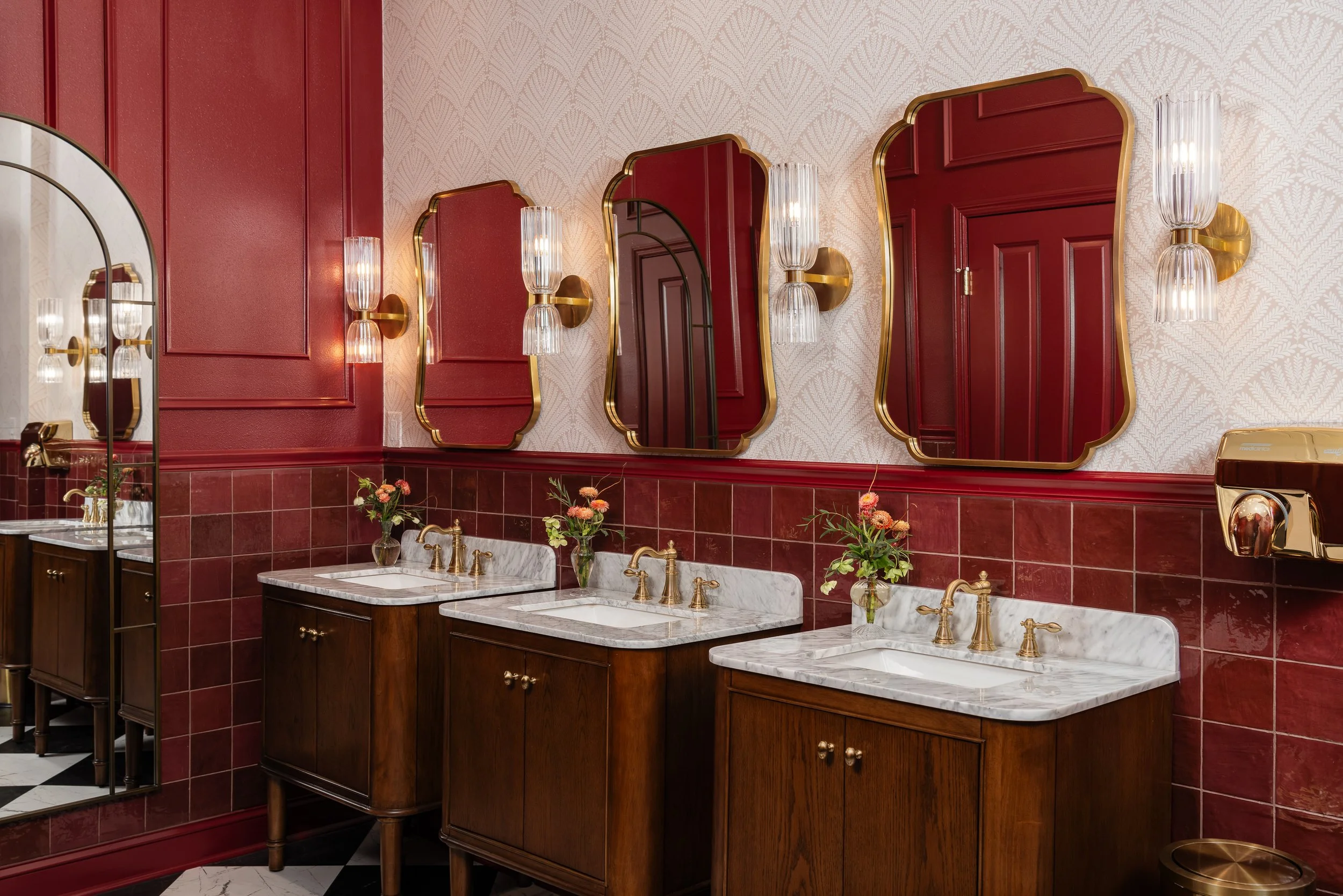 Three vanity sinks in a commercial restroom design with marble countertops and wooden cabinets are lined up against a red tiled wall, each with a unique gold-trimmed mirror and brass sconces above. Small vases of flowers sit on the sinks.