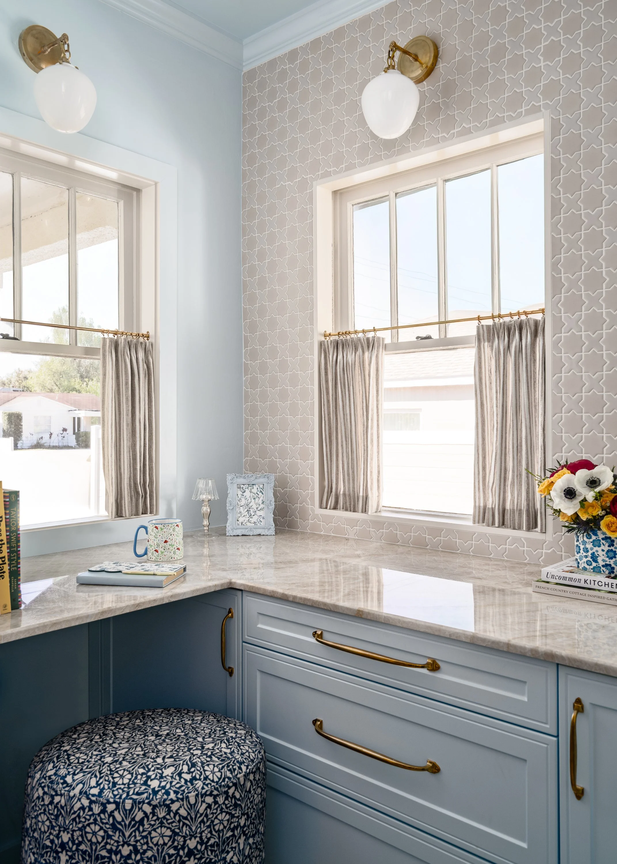 A light blue vanity with a marble countertop features two windows with beige curtains, brass sconces, and a patterned ottoman in the corner.