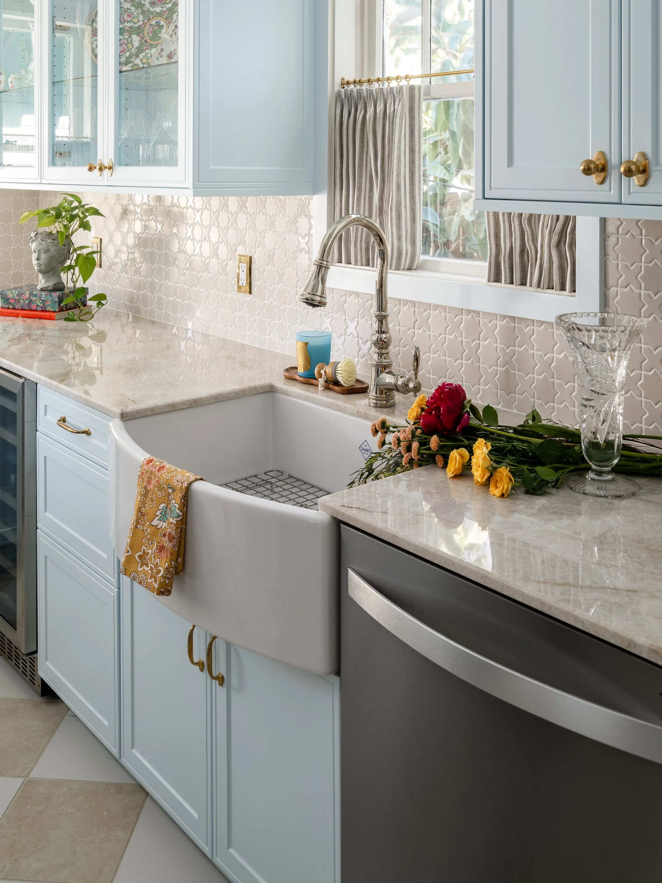 A bright kitchen sink area with light blue cabinets, a white apron sink, a polished nickel faucet, and a countertop decorated with flowers and a crystal vase.