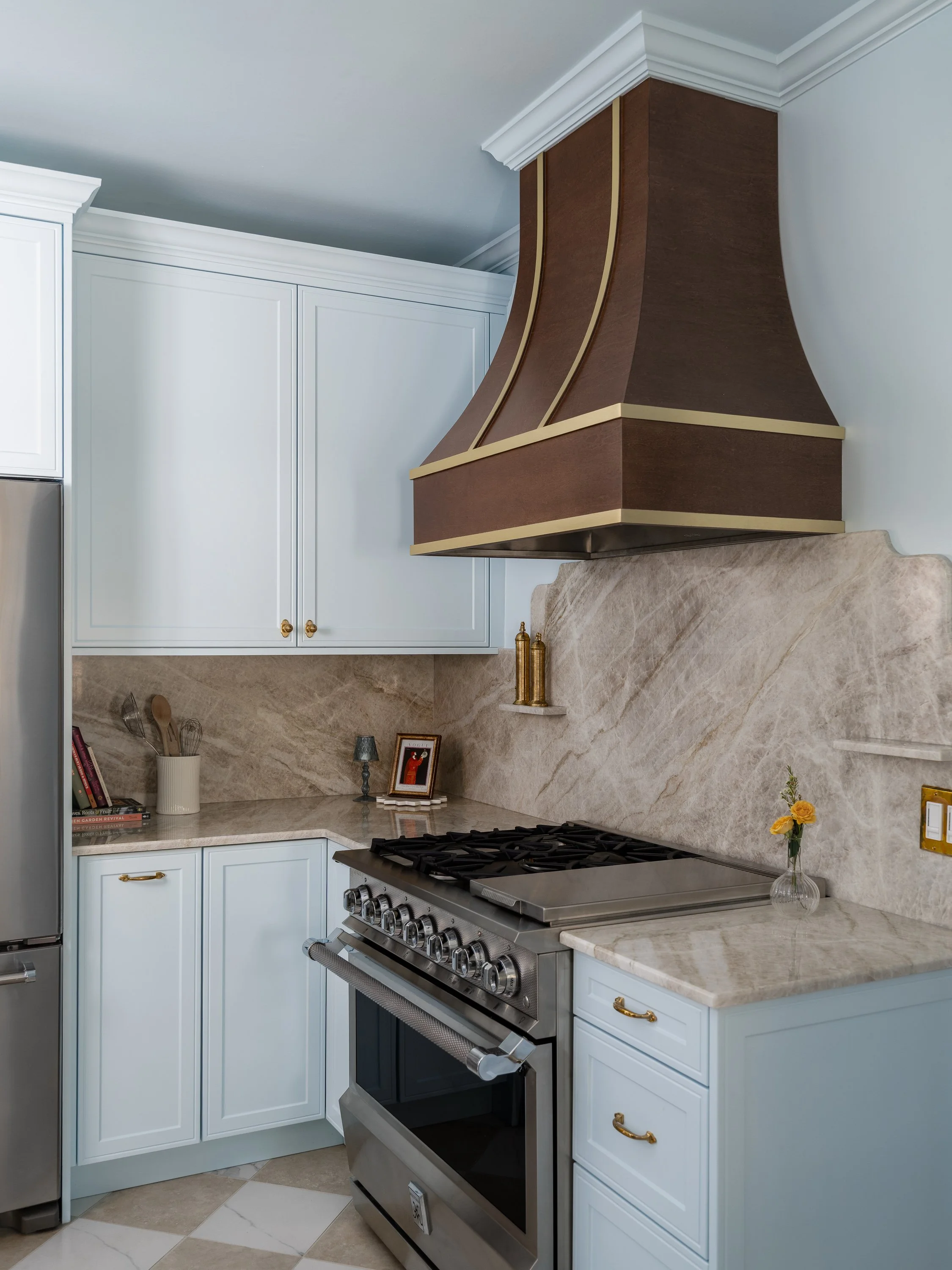 A kitchen with light blue cabinets, a brown and gold range hood, and a marble backsplash over a stainless steel range.