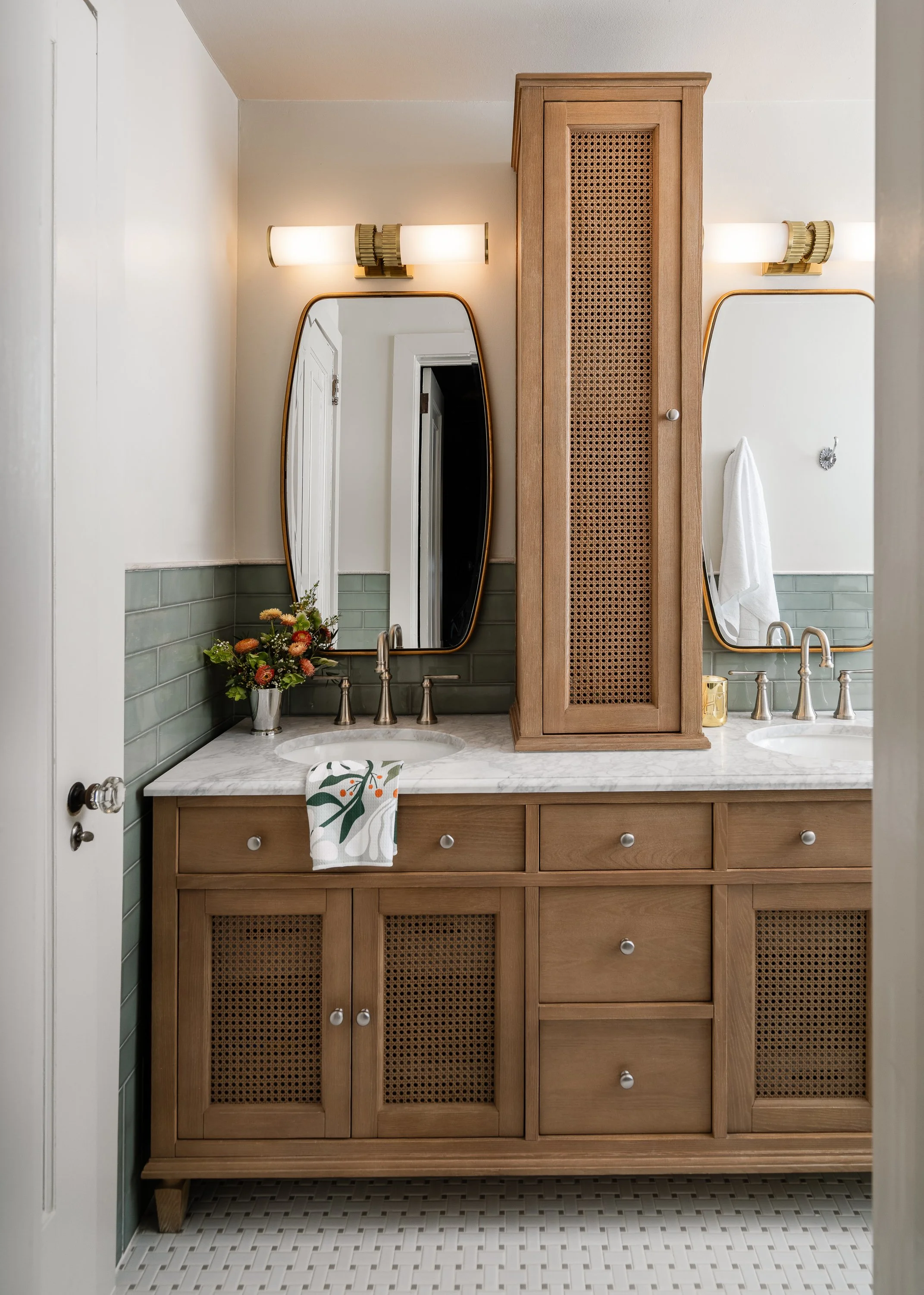 A wooden double vanity with cane detailing under two mirrors, each with brass sconces above. Green subway tile backsplash and a small vase of flowers complete the look.