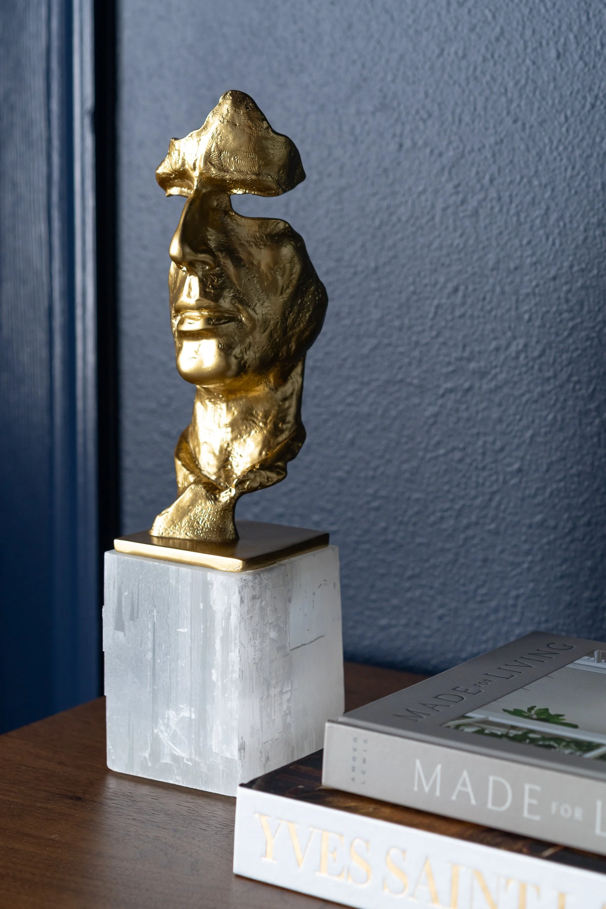 A golden bust of a face is mounted on a white selenite crystal base, placed on a wooden surface next to two books.