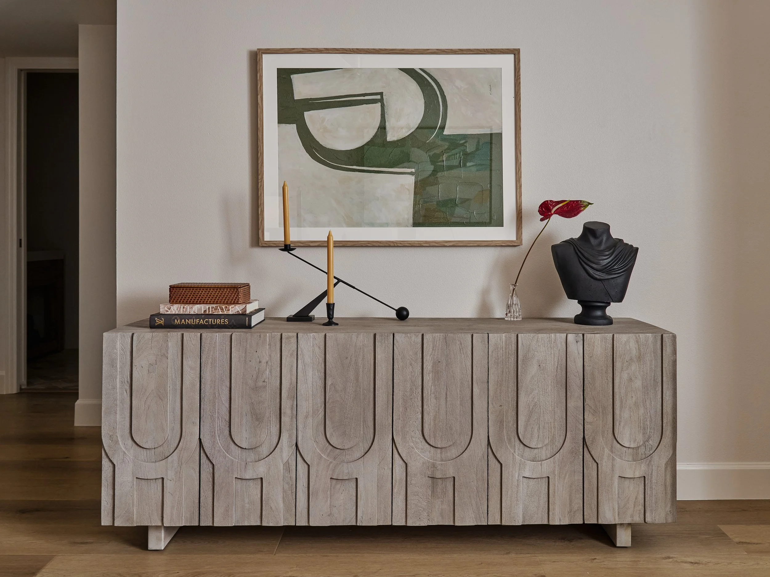 A living room wall with a framed abstract green-and-gray artwork above a light gray carved sideboard, styled with books, a small sculpture, a vase, and a red flower.