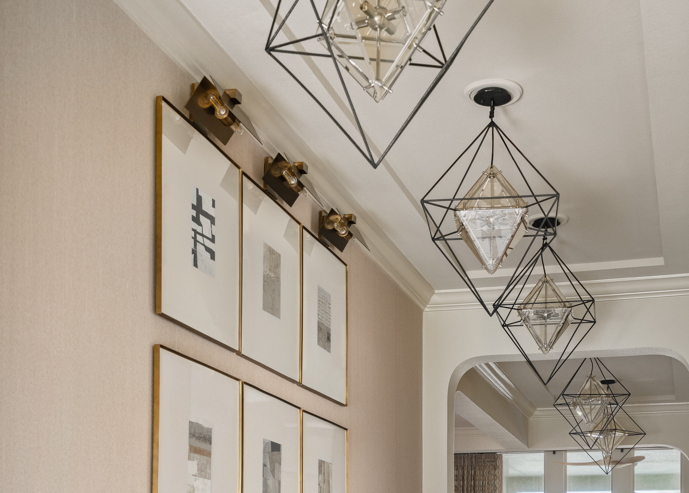 A row of framed artwork with gold trim hangs on a textured wall, beneath a series of modern geometric pendant lights.
