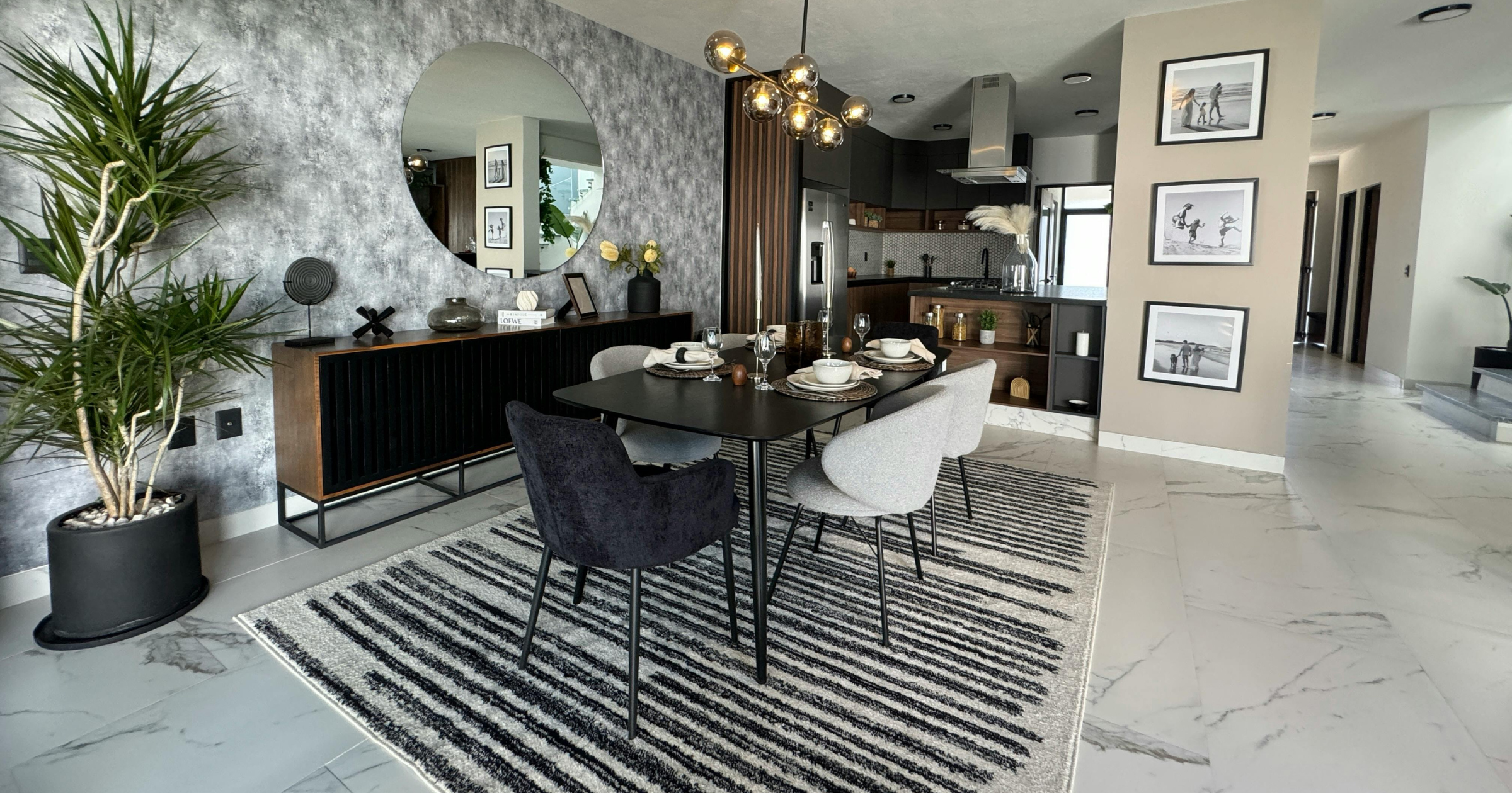Coastal dining room with textured wallpaper, black and white modern dining table and chairs, striped rug, marble floor, potted plant, modern kitchen in the background, family photos on the wall.