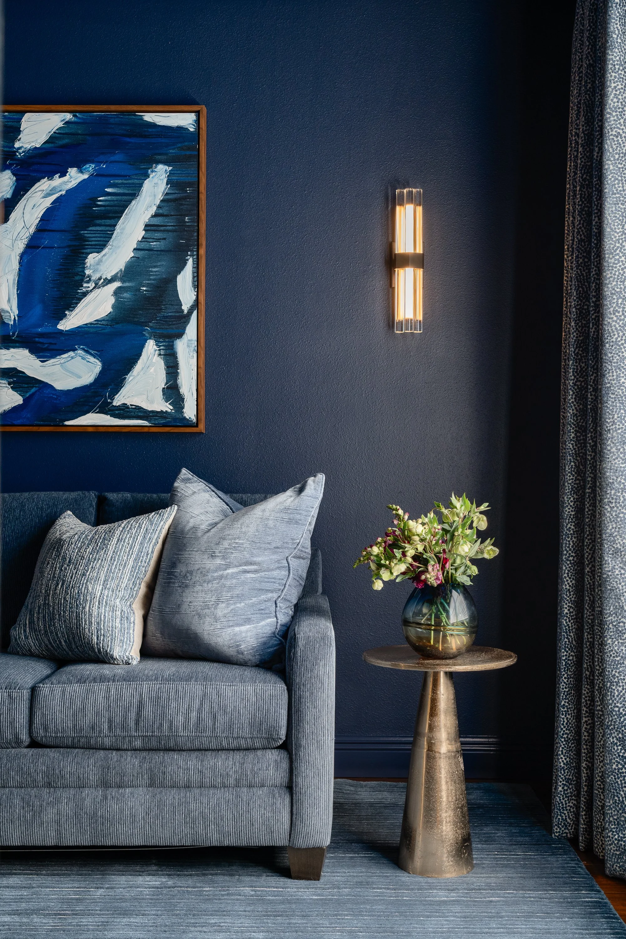 A contemporary modern sunroom design scene featuring a blue sofa with textured pillows, a metallic side table with a vase of flowers, a piece of abstract blue art on a dark blue wall, and a wall sconce.