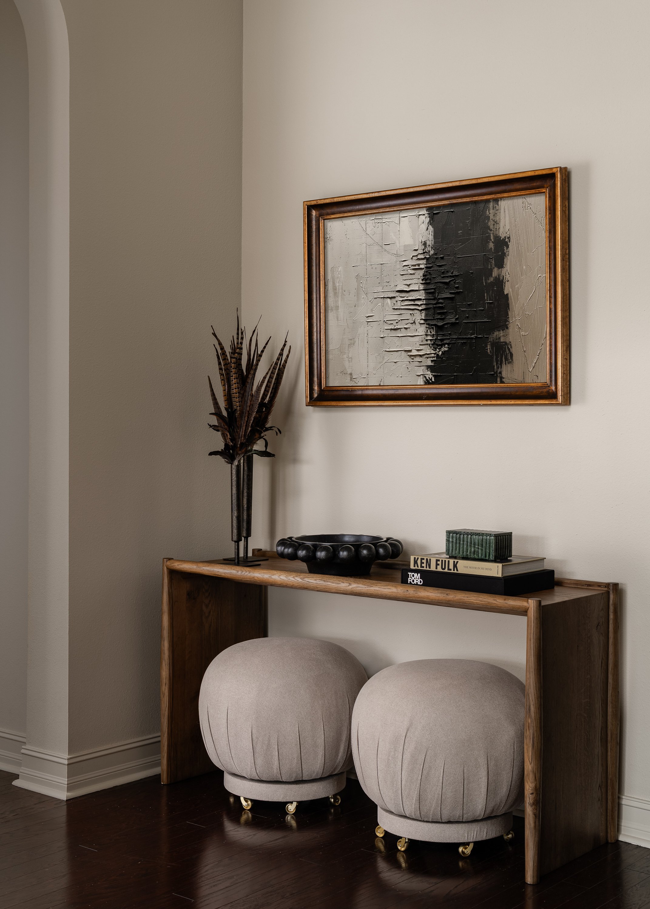 A wooden console table with a textured abstract painting above it and two round upholstered ottomans below. A vase with dried feathers and decorative objects sit on the table.