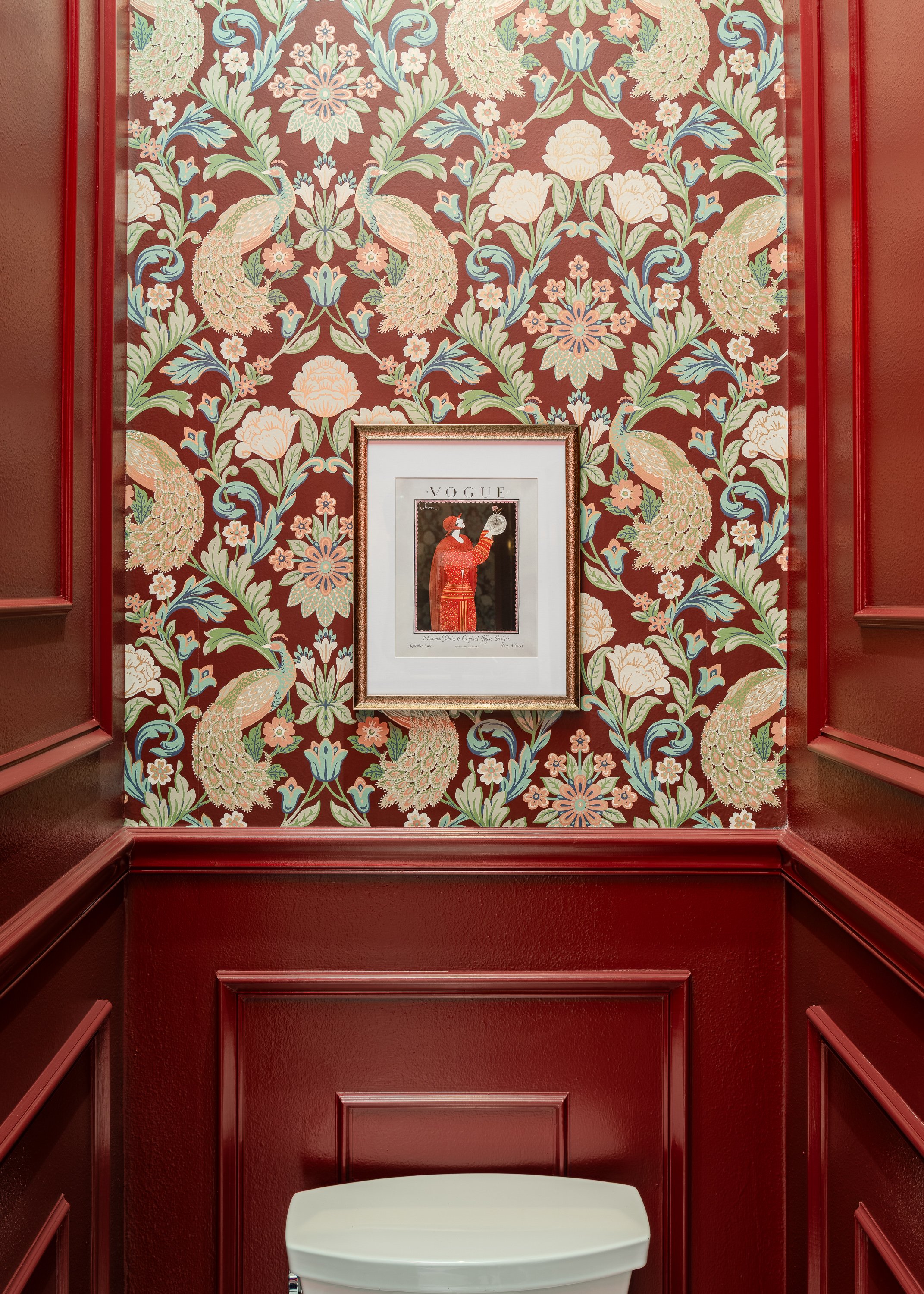 A commercial restroom design with a framed Vogue cover featuring a person in a red robe is displayed on a wall with red wainscoting and a bold peacock and floral wallpaper.