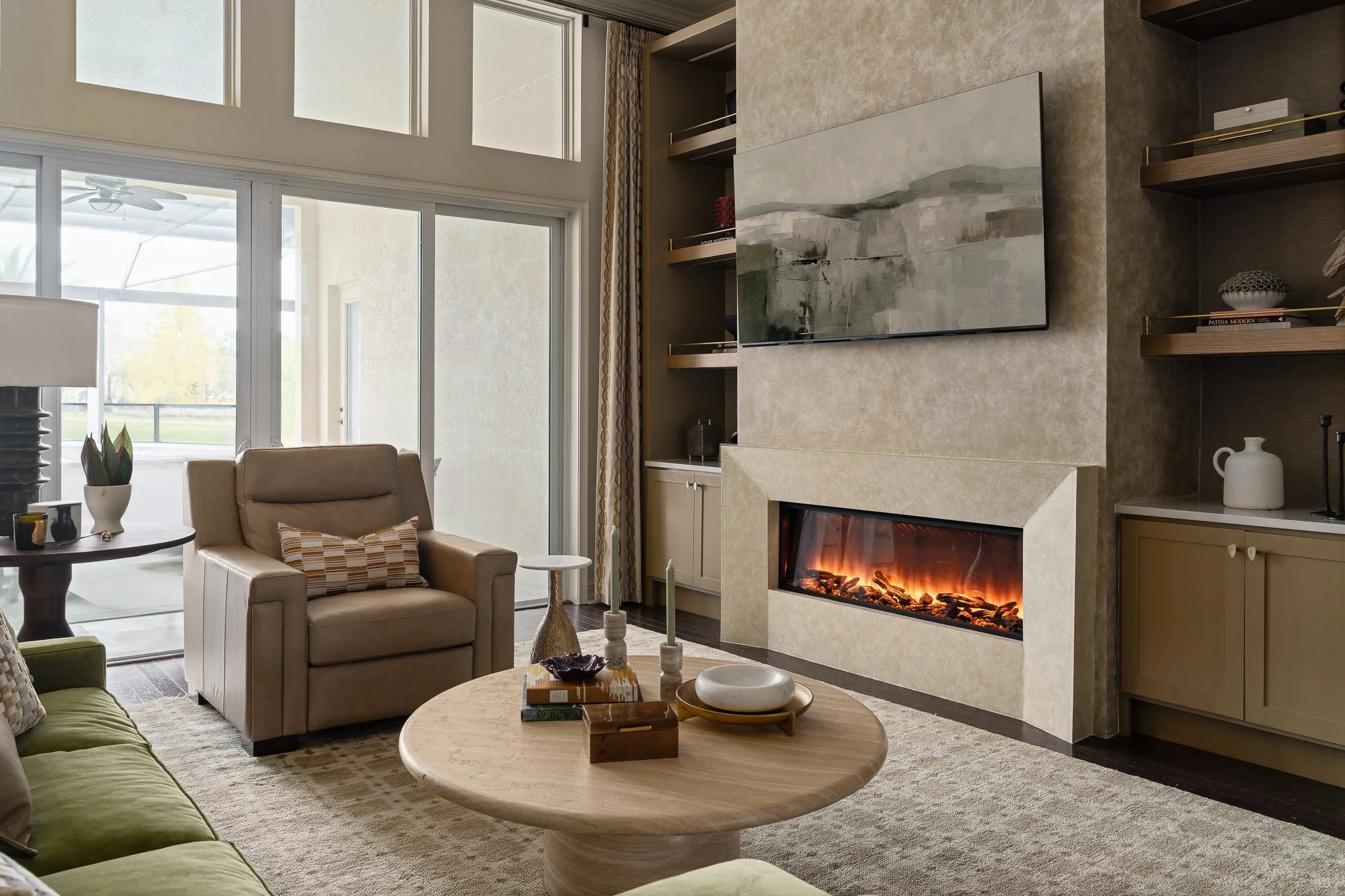 A living room features a plush tan armchair, a green sofa, and a round coffee table in front of a modern fireplace with a television mounted above it.