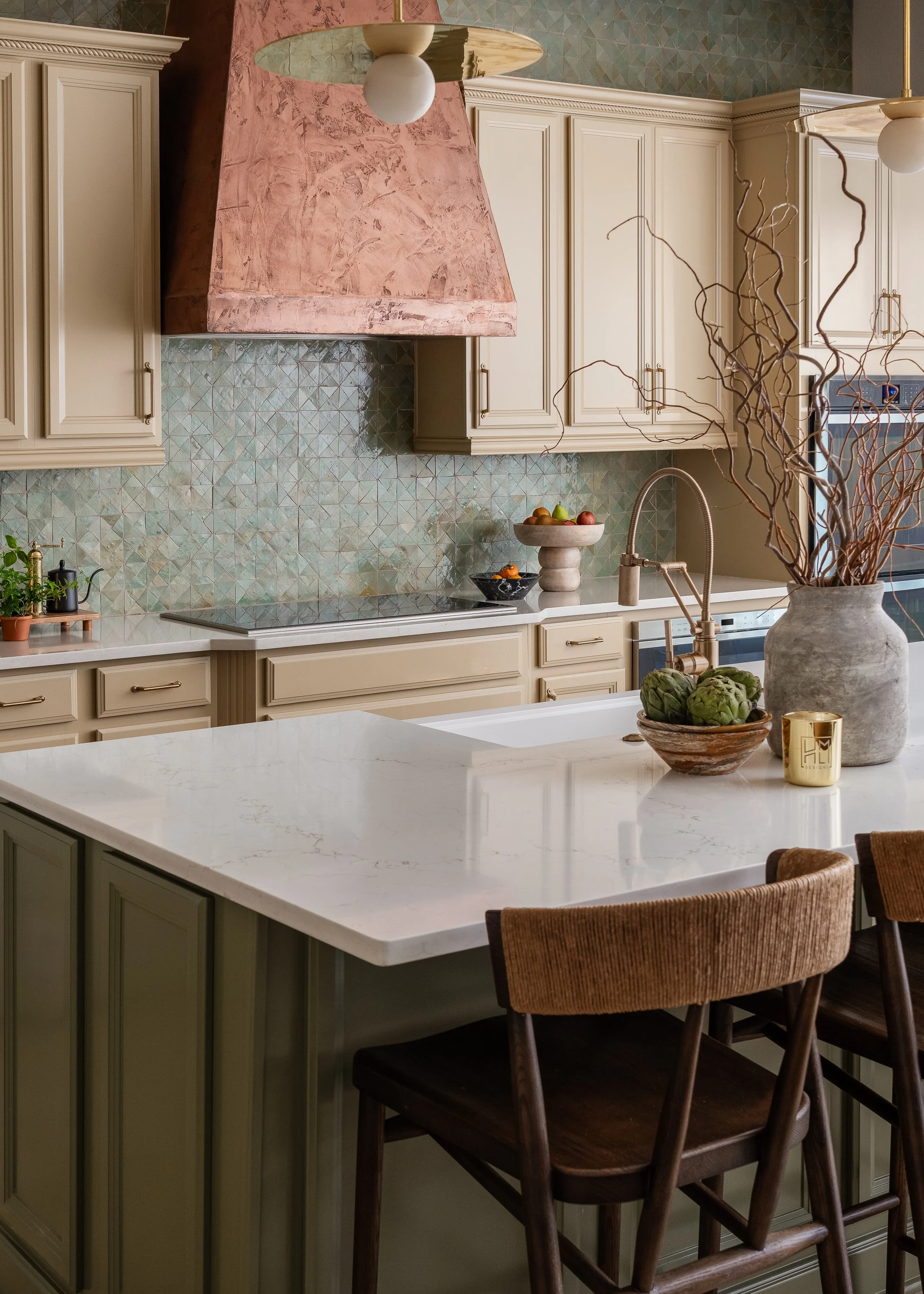 A modern kitchen features cream-colored cabinets, a copper range hood, and a backsplash with teal geometric tiles. A white quartz island with bar stools is in the foreground, with a vase of branches and a bowl of artichokes on the counter.