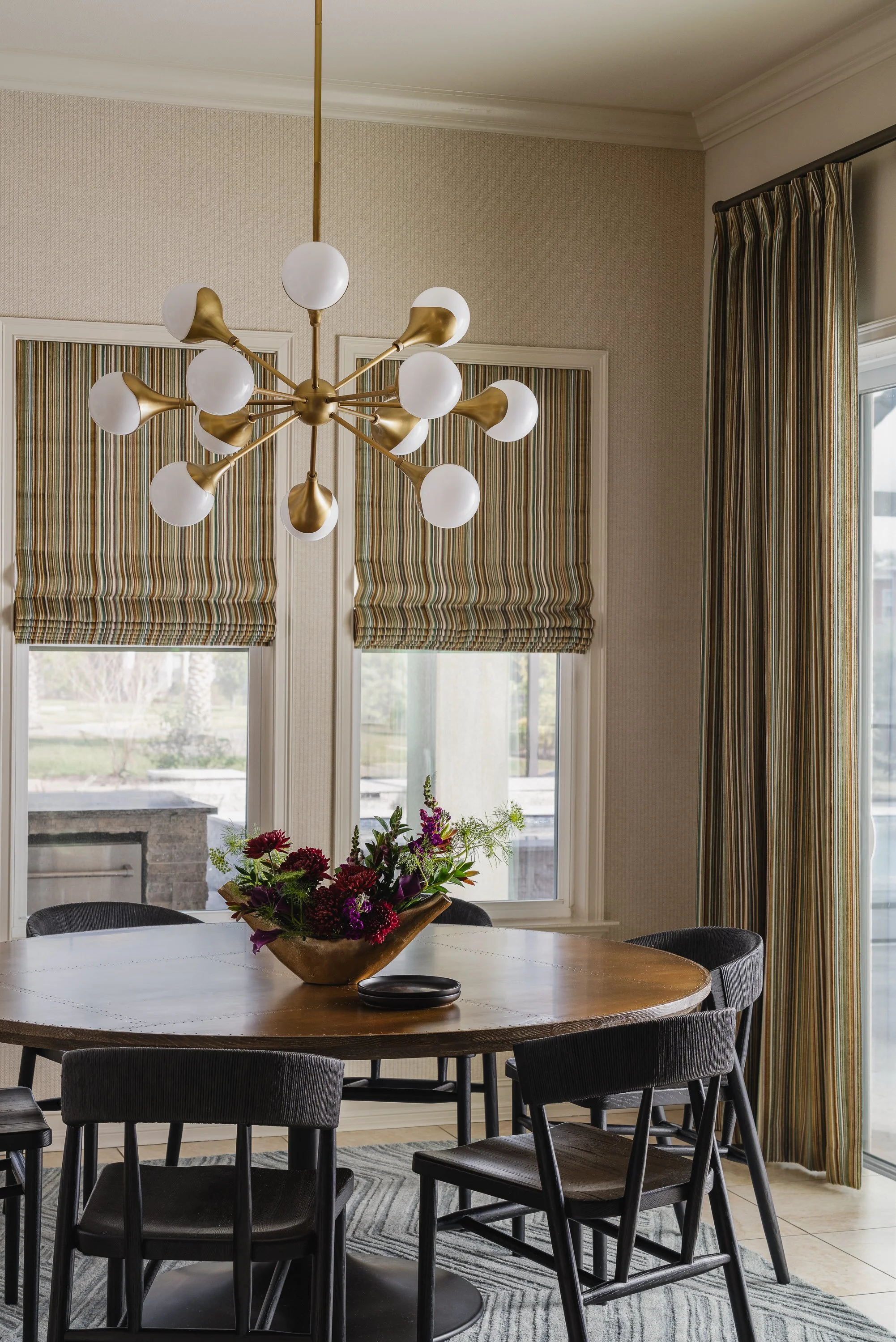 A round wooden dining table with dark chairs is set with a floral centerpiece under a Sputnik-style chandelier.
