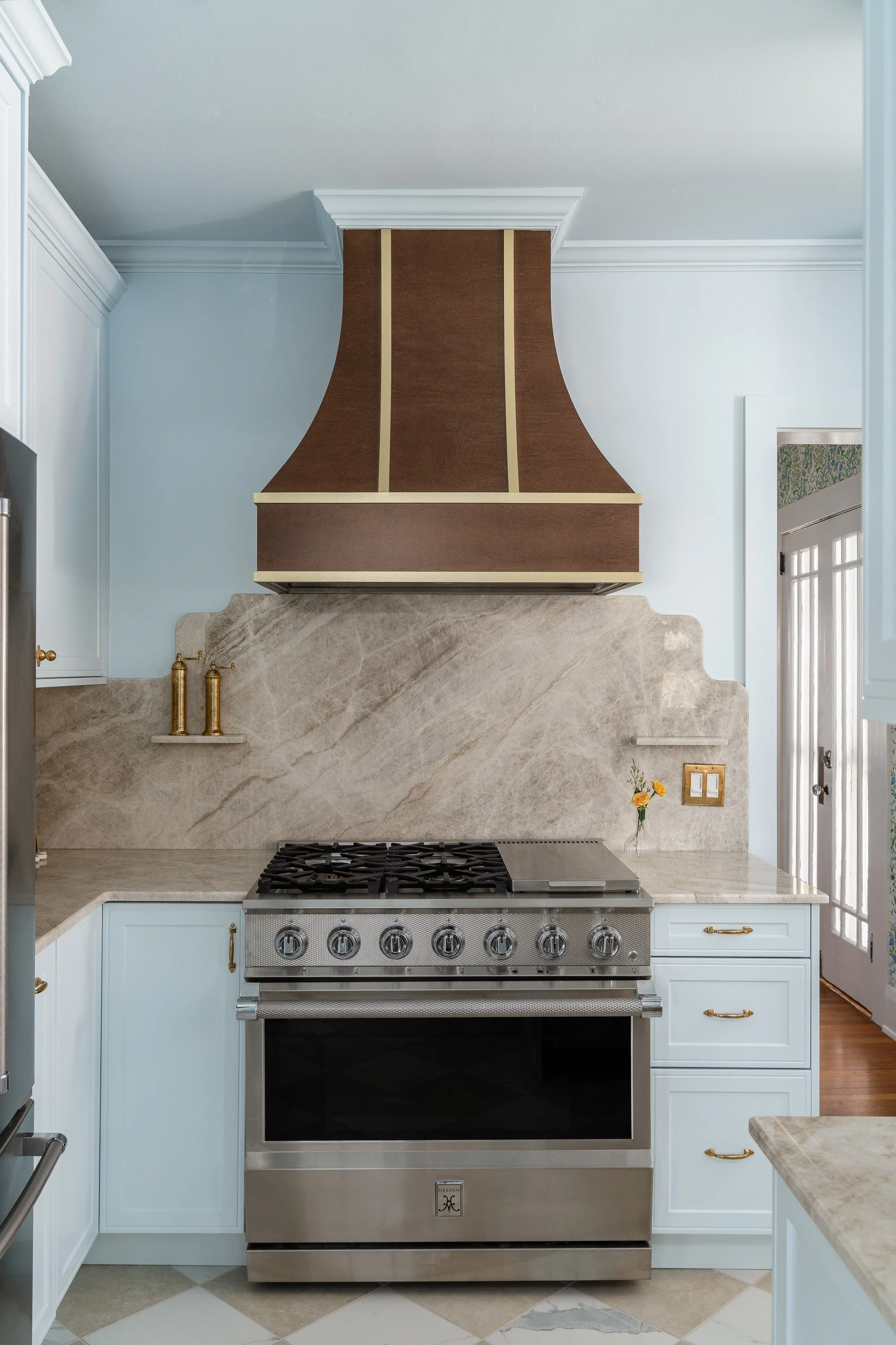 A stainless steel gas range is centered under a decorative brown and gold range hood with a marble backsplash. Light blue cabinets are on either side of the range.