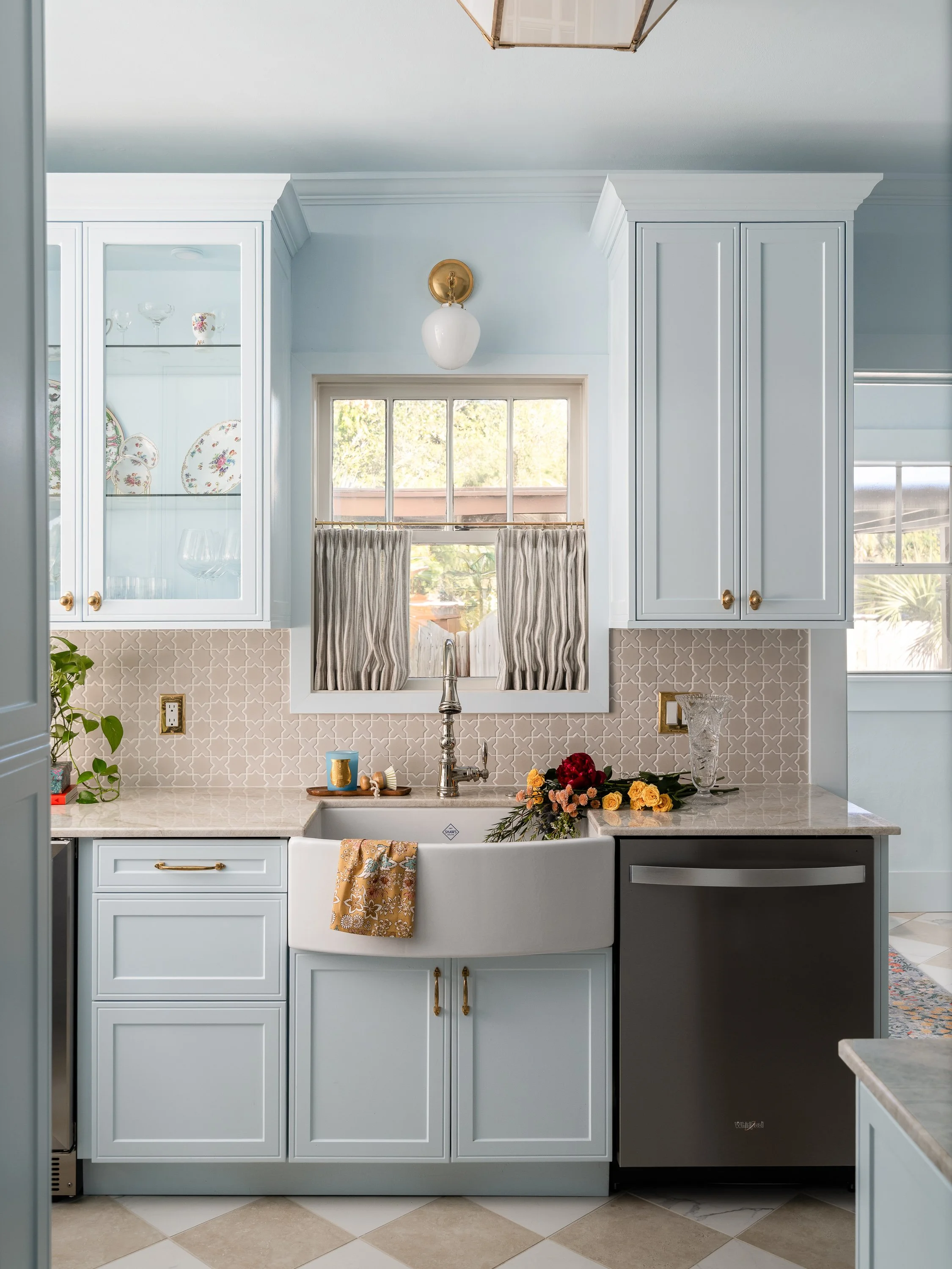 A light blue kitchen featuring a white farmhouse sink, a dark gray dishwasher, and a window with gray curtains above the sink. The backsplash is a beige tile with a repeating pattern, and the cabinets are light blue with gold hardware.