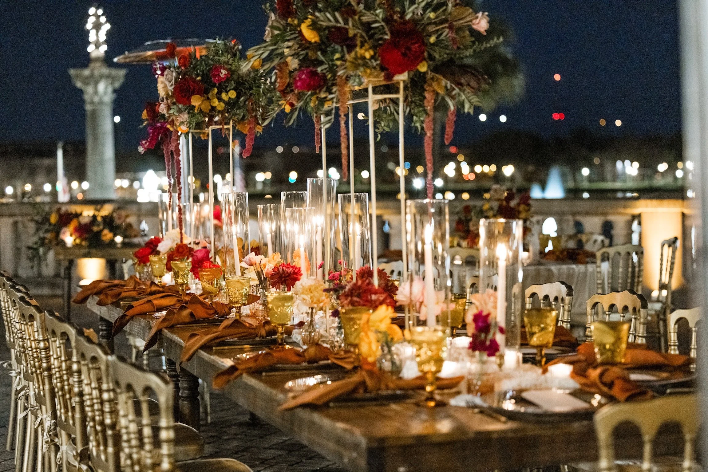 A long wooden table is set for a luxury wedding outdoors at night, decorated with tall floral centerpieces, candles in glass cylinders, and amber-colored glassware, and burgundy and champagne colored flowers. Rows of gold chairs are visible.