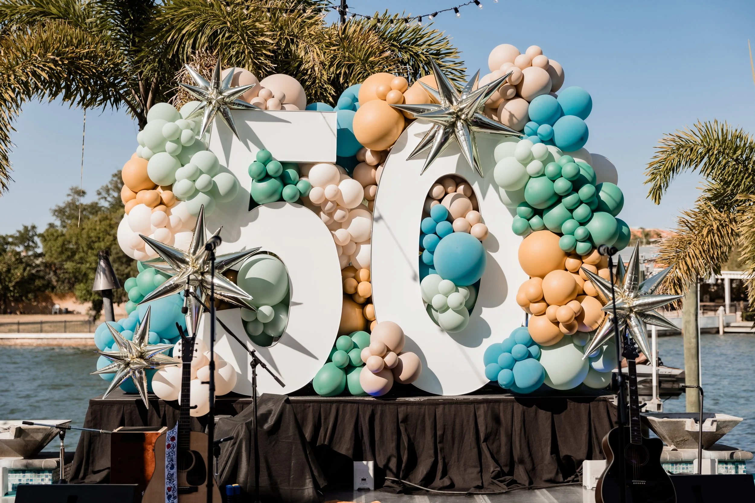 50th birthday party decor idea stage set up with giant number 50, teal, turquoise, green, and orange balloons, two guitars on a harbor