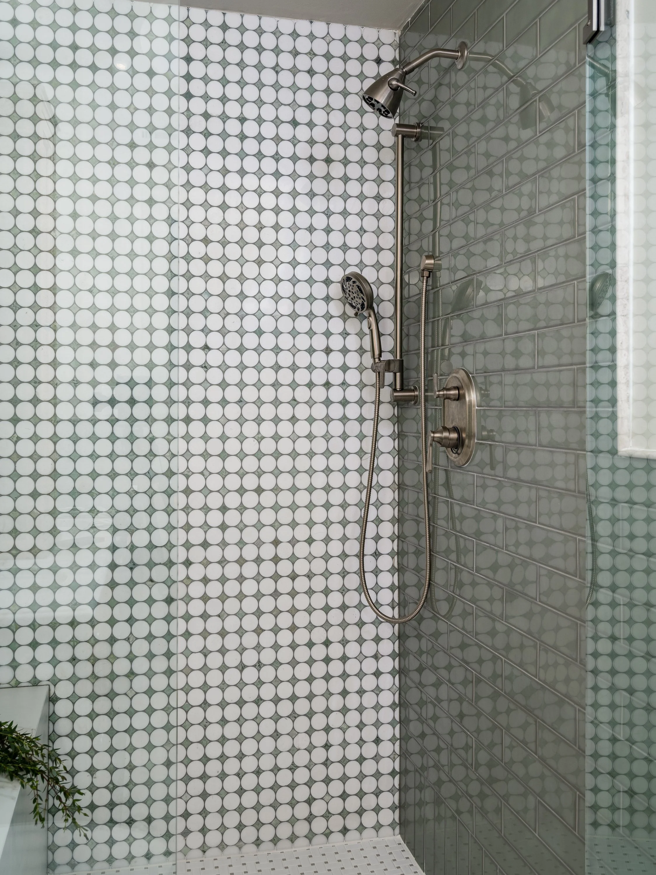 A modern shower with two shower heads and a glass enclosure, featuring penny tile on one wall and subway tile on the other.