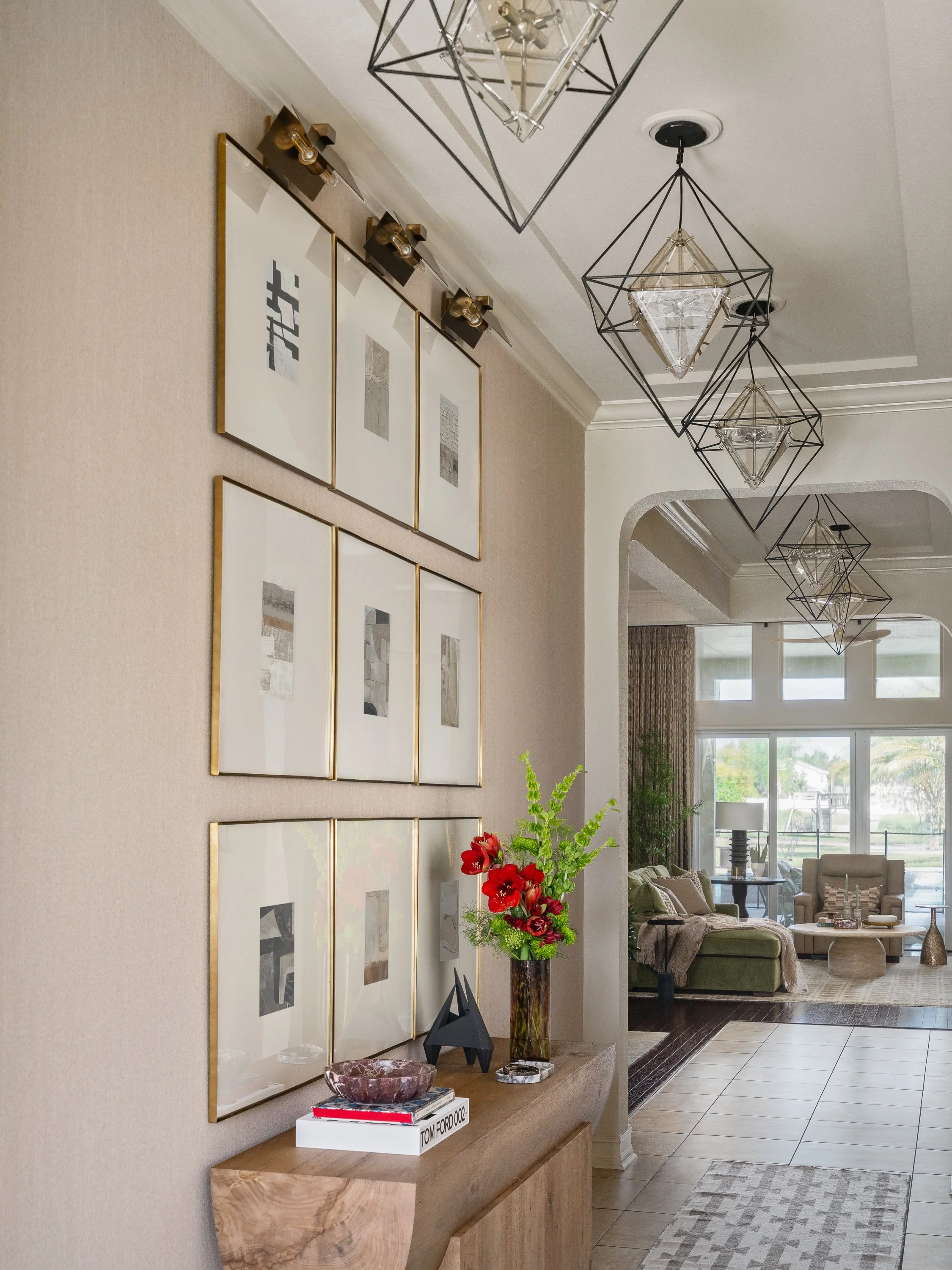 A hallway features a console table with a vase of red flowers and books, beneath a grid of framed art and geometric chandeliers.