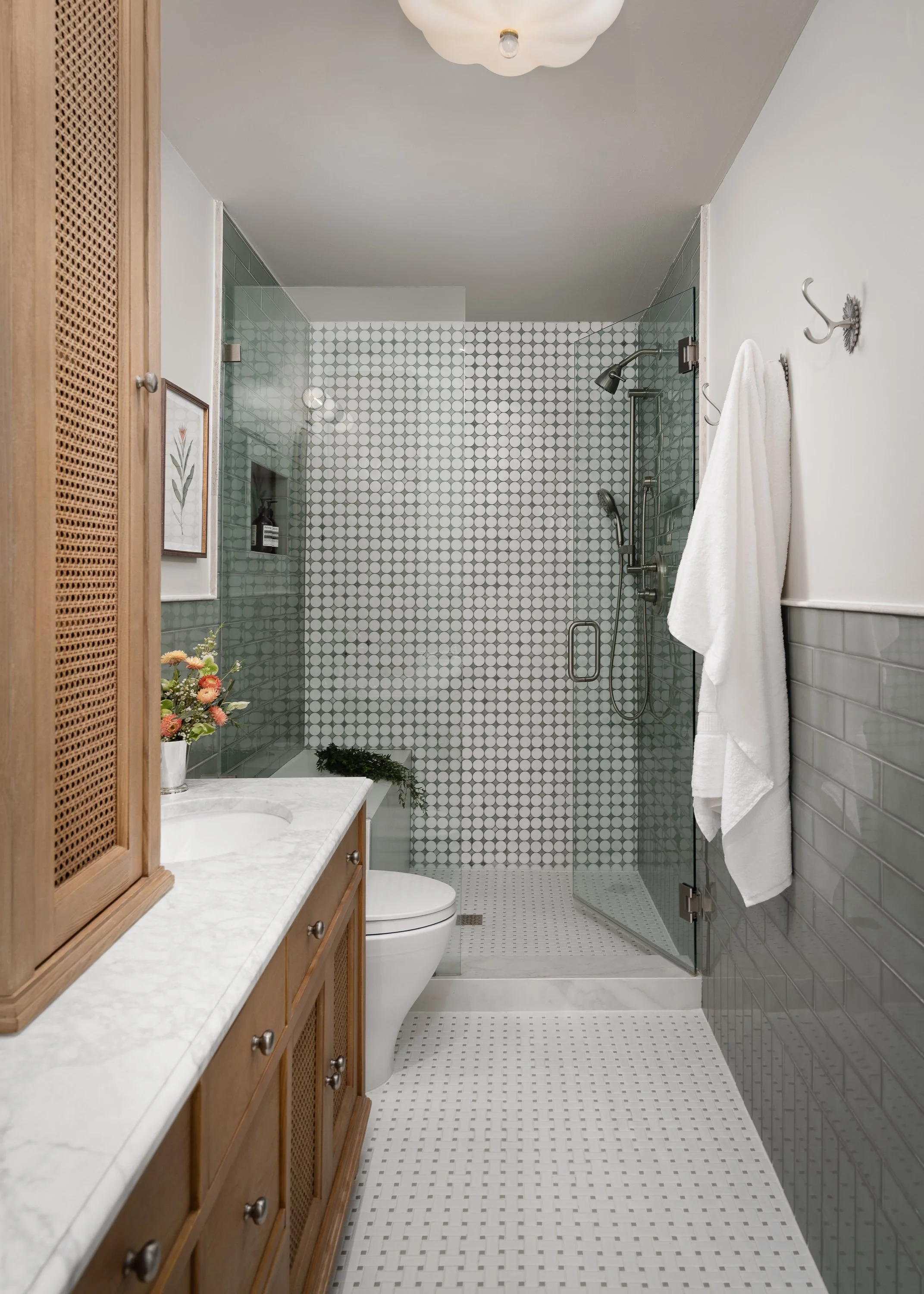 A modern bathroom features a wooden vanity with a marble countertop, a white toilet, and a glass-enclosed shower with patterned tile walls.