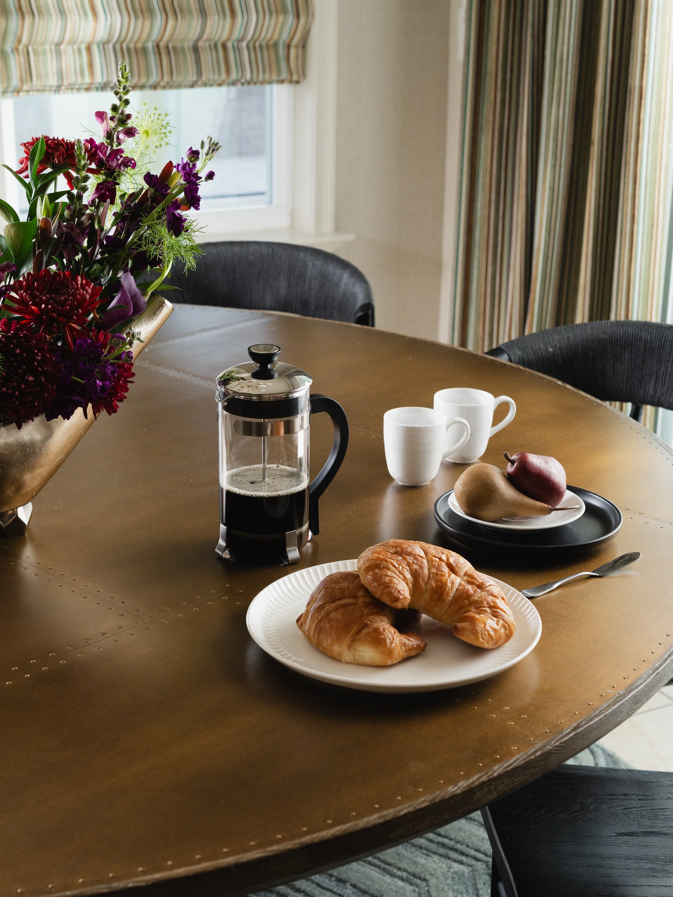 A French press coffee maker, two croissants on a plate, and two pears sit on a brass table next to a vase of flowers.