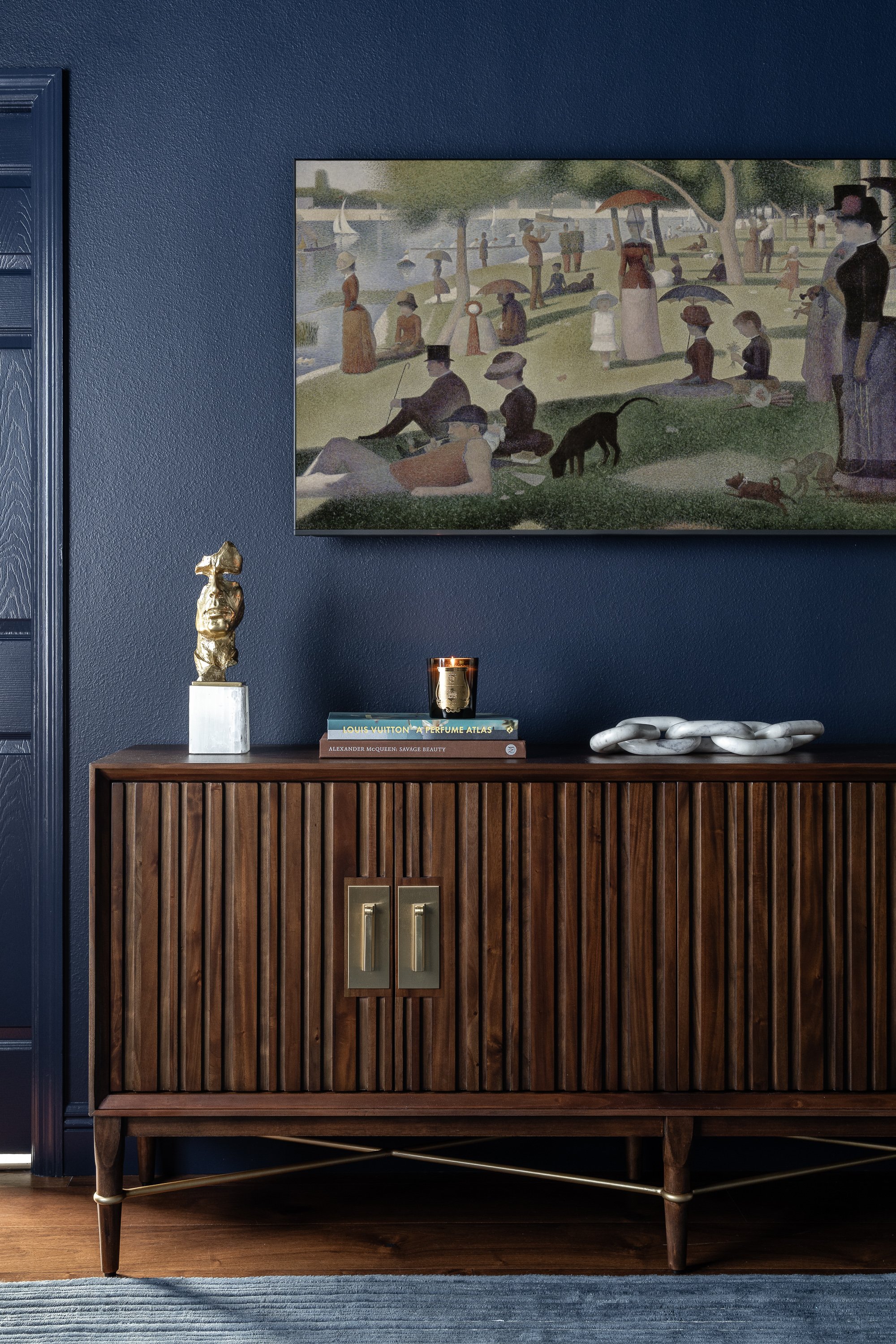 contemporary modern sunroom design. A dark wooden cabinet with fluted doors and brass handles is adorned with a gold sculpture, a candle, and books. Above, a painting depicts a lively park scene with people and a dog.